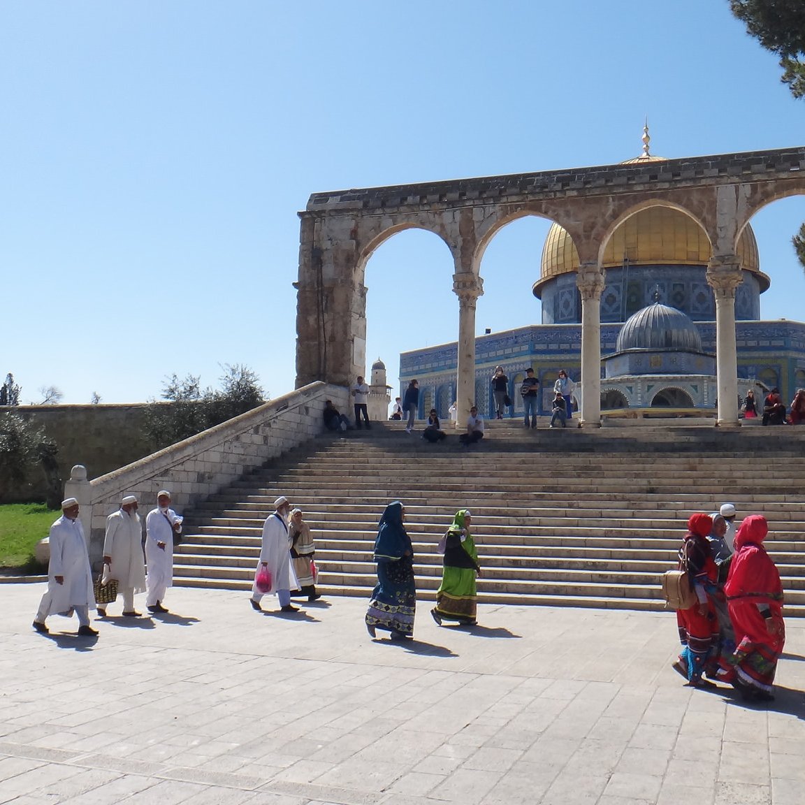 L'image montre un paysage architectural impressionnant avec des arches majestueuses en premier plan. On peut voir un groupe de personnes marchant sur une large marche, certaines portant des vêtements traditionnels colorés. En arrière-plan, la célèbre mosquée avec son dôme doré est visible, entourée d'un ciel clair et d'arbres verdoyants. L'atmosphère semble paisible et spirituelle, reflet d'un lieu de rassemblement important.
