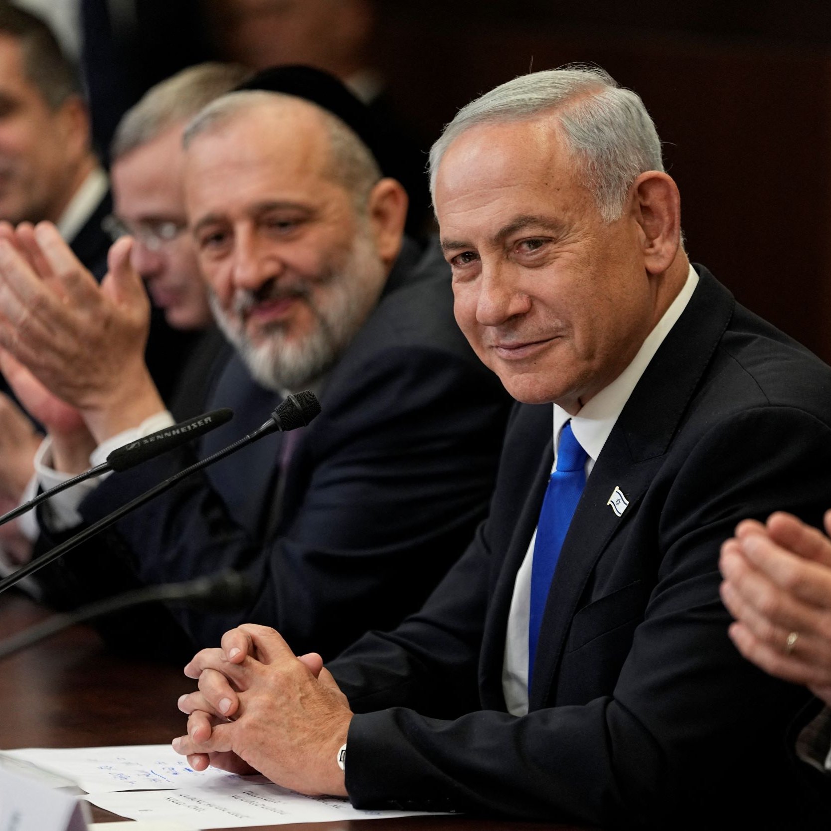 تظهر الصورة مجموعة من الرجال يجلسون حول طاولة اجتماع. في المقدمة، يوجد رجل يرتدي بدلة سوداء وقميص أبيض، يظهر على وجهه تعبير هادئ ويبتسم. إلى جانبه، يجلس رجل آخر يبدو سعيدًا ويصفق، بينما يظهر رجال آخرون في الخلفية يبدون مشغولين بالحديث أو التصفيق. الخلفية تحتوي على ملامح من قاعة اجتماعات.