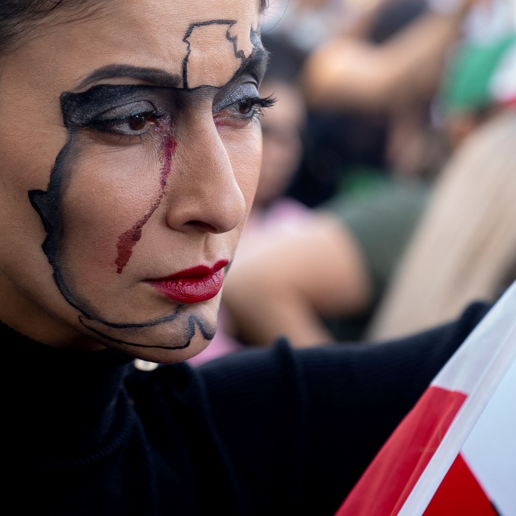 L'immagine mostra una donna con un trucco facciale elaborato, che rappresenta una mappa del suo paese. Ha un'espressione seria e intensa, con un tratto rosso che scende lungo il suo viso, suggerendo emozione o una narrazione profonda. In una mano tiene una bandiera, evidenziando un forte senso di identità nazionale e di protesta. Sullo sfondo si intravedono altre persone, creando un'atmosfera di raduno o manifestazione.