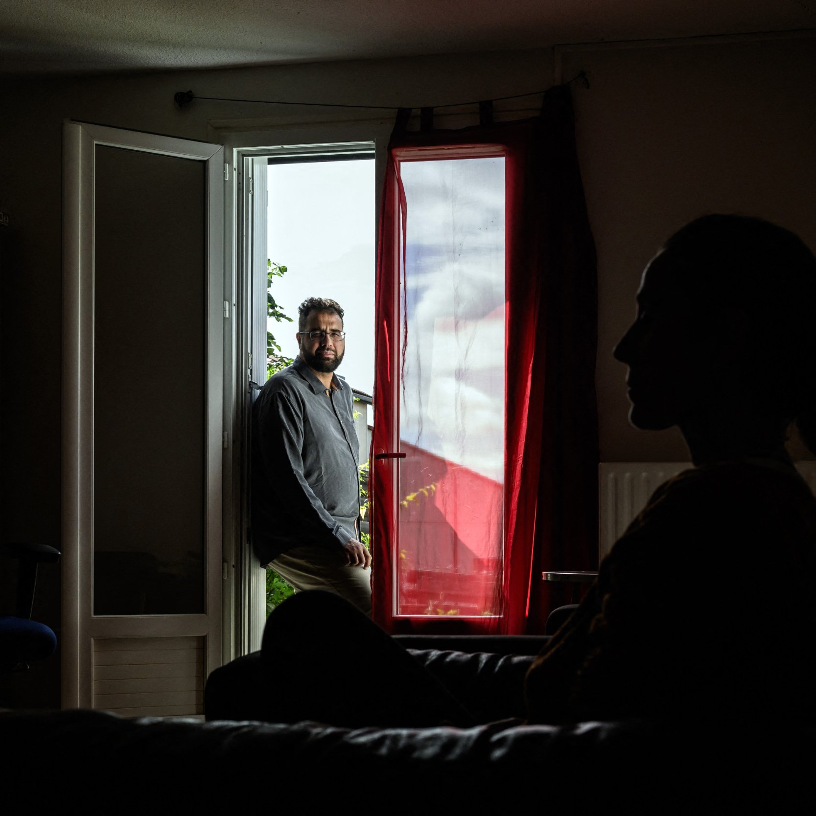 L'image montre un intérieur de maison ou d'appartement. Au premier plan, on voit la silhouette d'une personne assise sur un canapé, éclairée par une lumière douce, tandis qu'en arrière-plan, un homme est debout près d'une porte ouverte qui donne sur l'extérieur. La lumière entre par la fenêtre de façon à créer un contraste intéressant entre les deux personnages. Les éléments de la pièce, tels qu'un fauteuil et des étagères, ajoutent à l'ambiance chaleureuse et intime de la scène.