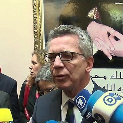 تُظهر الصورة شخصًا يتحدث أمام مجموعة من الكاميرات والميكروفونات، ويبدو أنه في مؤتمر صحفي أو حدث رسمي. خلفه، يوجد صورة كبيرة ليد مع بعض الرموز، ربما تتعلق بالتراث أو الثقافة. هناك أيضًا عدد من الأشخاص الآخرين يقفون في الخلفية، مما يشير إلى أهمية الحدث أو اللقاء. الأعلام التي تظهر في الصورة تعزز الطابع الرسمي للموقف.