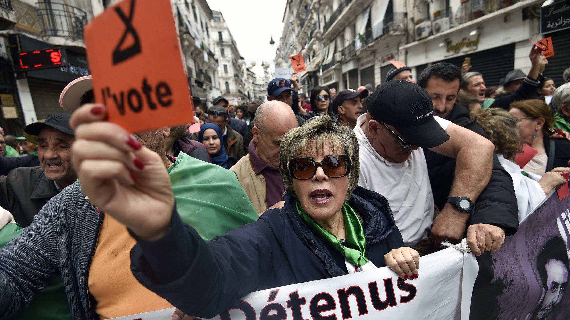 L'image montre une manifestation dans une rue d'Alger. Au premier plan, une femme tient une pancarte orange avec le mot "l'vote". Elle est entourée par d'autres manifestants, dont certains portent des drapeaux algériens. L'atmosphère semble engagée et déterminée, avec des expressions de solidarité parmi les participants. Des banderoles sont visibles, indiquant des revendications liées à la situation politique. L'environnement urbain et les bâtiments en arrière-plan témoignent d'une ambiance dynamique de protestation.