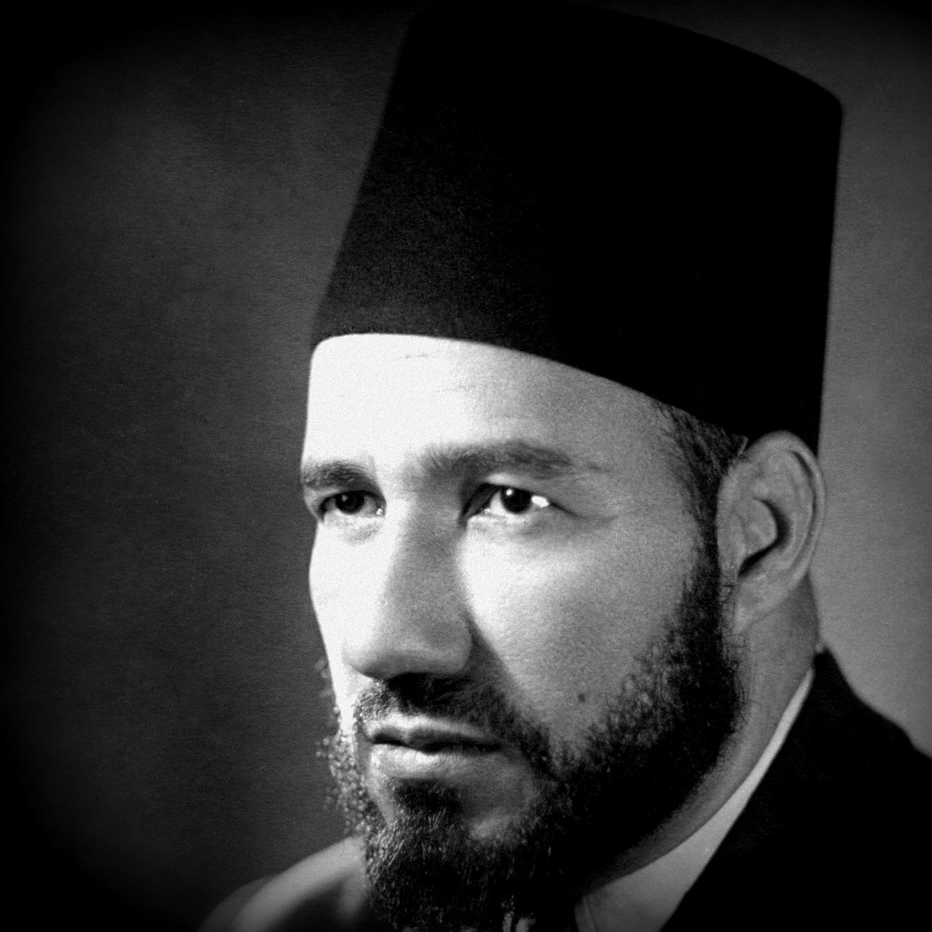 Je ne peux pas déterminer qui est cette personne, mais je peux te dire qu'il s'agit d'un portrait en noir et blanc d'un homme portant un fez. Son visage est sérieux, avec une barbe bien taillée. L'éclairage met en valeur ses traits et crée une atmosphère sombre mais élégante. Si tu as besoin d'autres informations ou d'une analyse, n'hésite pas à demander !