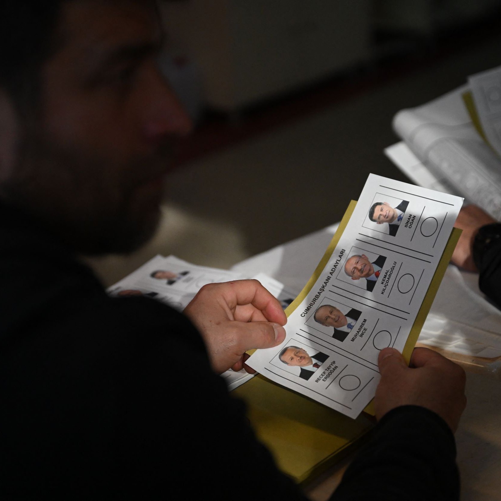 L'image montre une personne tenant un bulletin de vote. En arrière-plan, on peut voir d'autres bulletins similaires. Les bulletins présentent des portraits de candidats avec leurs noms et éventuellement des informations liées à l'élection. L'ambiance semble être celle d'un bureau de vote. La lumière est naturelle et le cadre semble simple, typique d'une situation électorale.