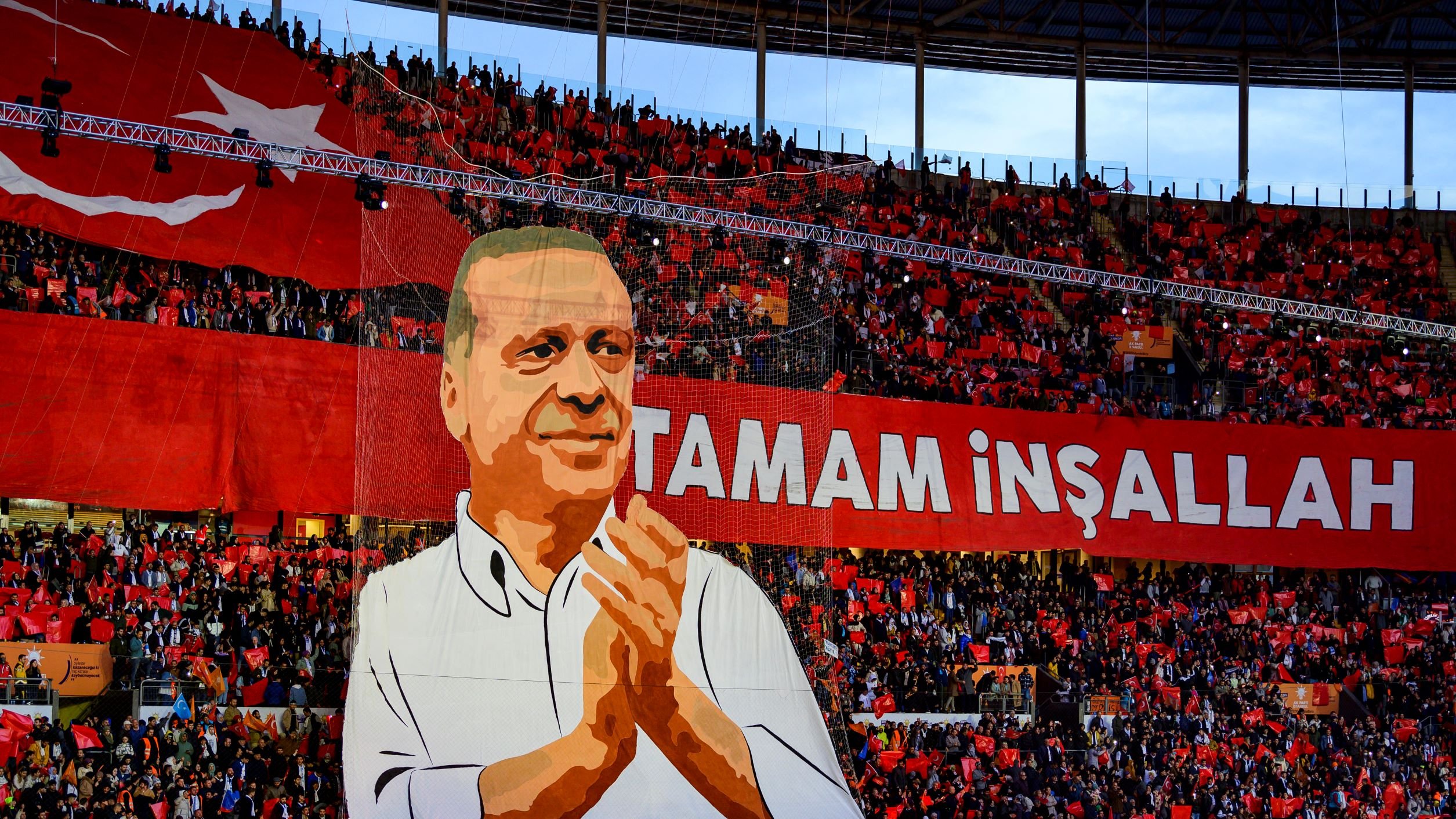 The image depicts a large crowd in a stadium, with a significant banner displayed prominently. The banner features a representation of a person, possibly a political figure, who is shown applauding. Surrounding the figure are enthusiastic supporters holding a sea of red flags or banners. The text "TAMAM İNŞALLAH" is written in bold letters, which translates to "It will be done, God willing" in English, conveying a message of hope or determination. The overall atmosphere appears vibrant and charged with energy, indicative of a political rally or celebration.
