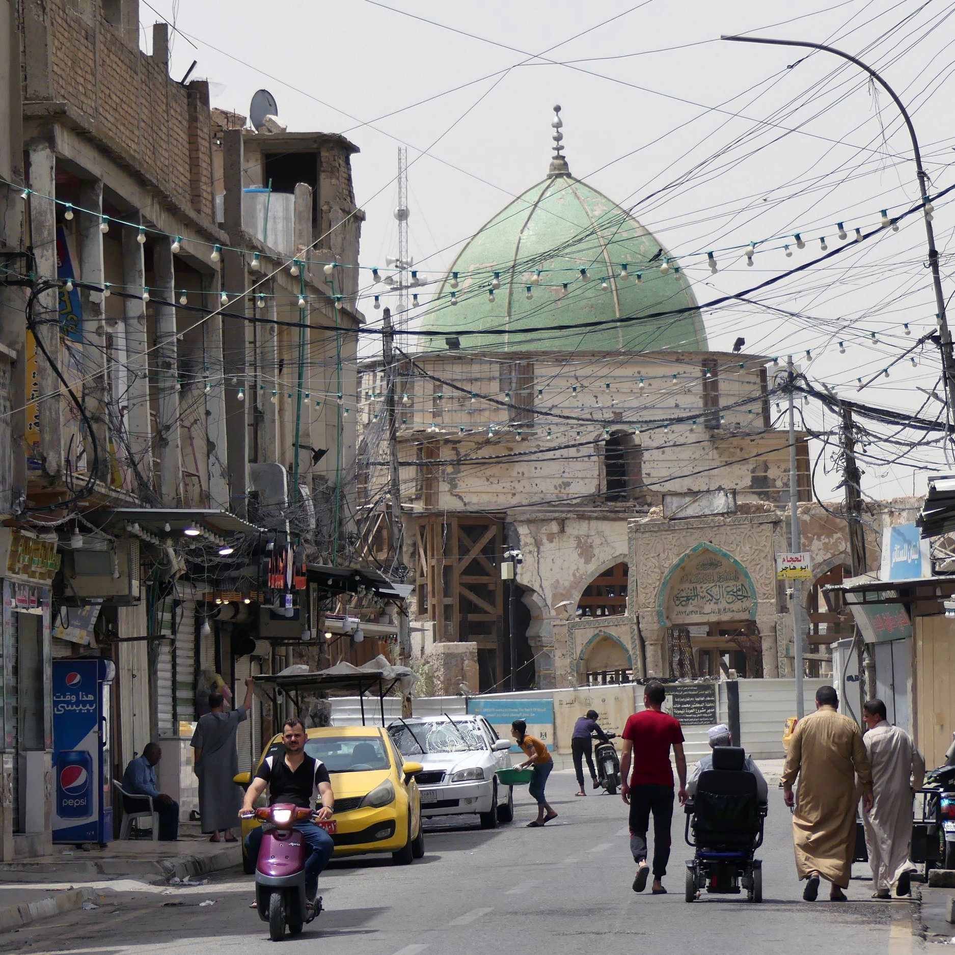 L'image montre une rue animée dans une ville. On y voit des bâtiments anciens avec des façades en pierre, certains étant en cours de rénovation. Au fond, une grande coupole verte domine la scène, indiquant la présence d'un bâtiment religieux. Les fils électriques sont visibles au-dessus de la rue. Des personnes marchent sur le trottoir, et une moto ainsi qu'une voiture jaune circulent. L'atmosphère semble à la fois traditionnelle et dynamique.
