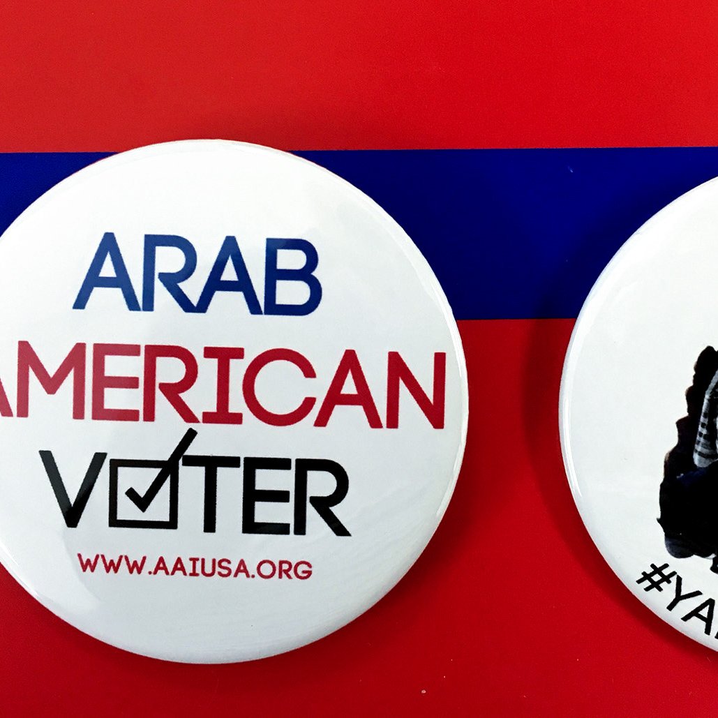 الصورة تحتوي على زرين دائرين. الزر الأول مكتوب عليه "ARAB AMERICAN VOTER" مع علامة التحقق، مما يدل على التصويت للمواطنين العرب الأمريكيين. الزر الثاني يظهر صورة لشخص يرتدي قبعة تحمل نجمة، وهو رمز معروف في الثقافة الأمريكية، مع عبارة "#YALLAVOTE" التي تدعو الناس للتصويت. الخلفية حمراء مع شريط أزرق، مما يضيف طابعاً احتفالياً للموضوع.