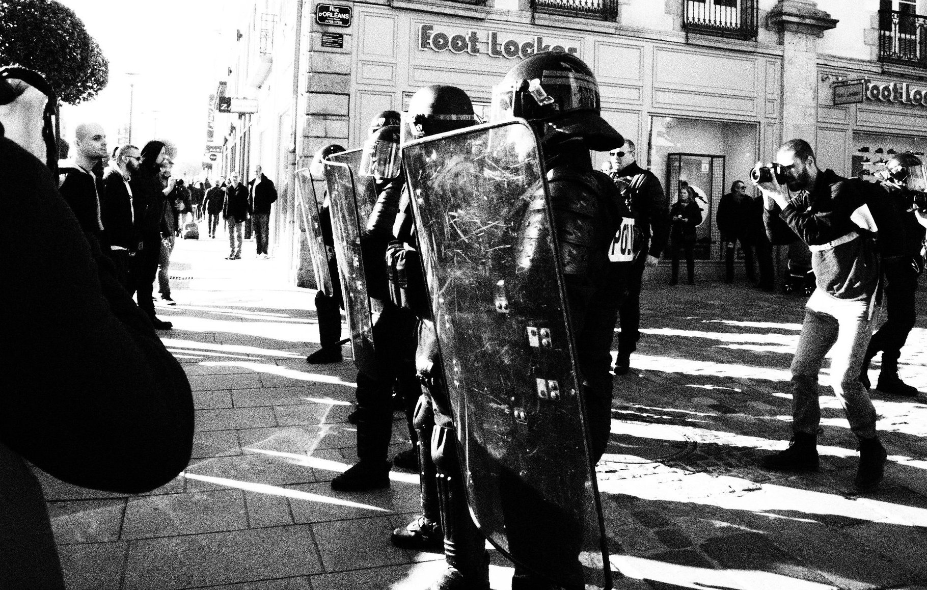 L'image montre une scène urbaine en noir et blanc, où des policiers en tenue anti-émeute, portant des casques et tenant des boucliers, se tiennent en position sur une rue. Ils sont face à une foule de personnes, dont certaines semblent prendre des photos. L'environnement est composé de bâtiments commerciaux, et l'ambiance semble tendue. Les ombres et la lumière créent un contraste saisissant dans la composition.
