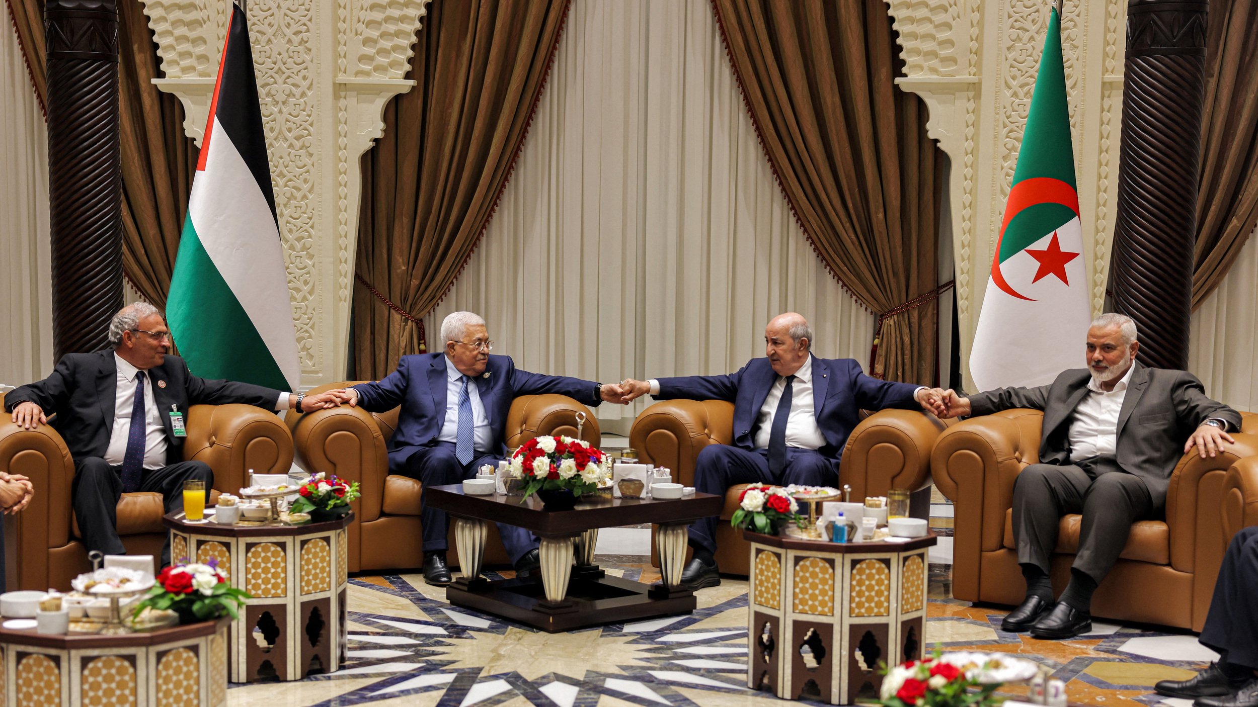 تظهر الصورة مجموعة من الأشخاص seated في غرفة مزينة بشكل أنيق، حيث يجلس أربعة رجال على أرائك مريحة. بينهم أجواء من التعاون والود، إذ يتبادلون الحديث مع قيامهم بتشابك الأيادي. في الخلفية، توجد أعلام فلسطين والجزائر، ما يشير إلى وجود موضوع يتعلق بالتعاون العربي أو القضايا السياسية بين البلدين. الطاولات أمامهم مزينة بتنسيقات من الزهور ومشروبات. الأجواء تبدو رسمية ولكن ودية في الوقت نفسه.