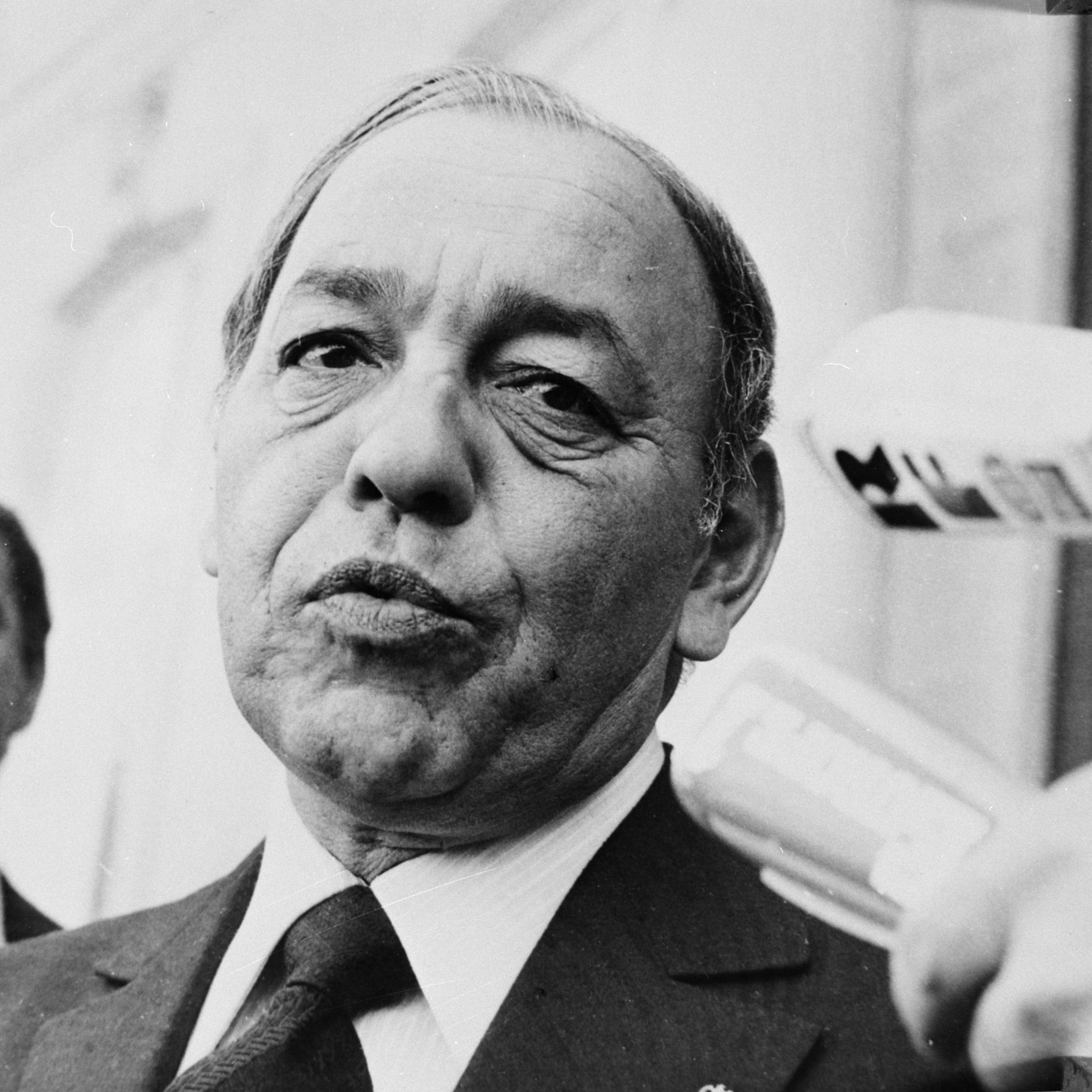 L'image montre un homme en costume, avec une expression sérieuse, s'adressant à des journalistes ou à une caméra. Il est situé au premier plan, tandis qu'un homme en arrière-plan semble être un gardien de sécurité ou un garde du corps. L'environnement semble être extérieur, probablement à un événement médiatique. Les microphones pointés vers lui indiquent qu'il donne une déclaration ou répond à des questions.