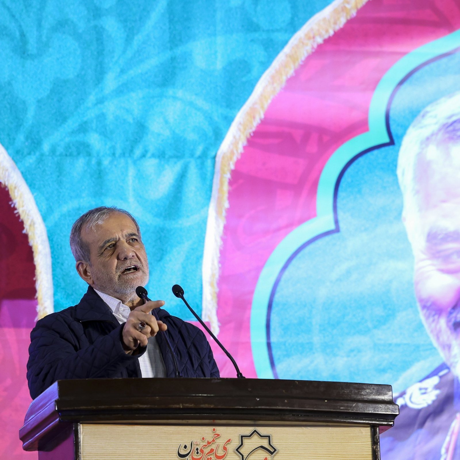 The image features a man speaking at a podium, with a colorful backdrop that includes a large portrait of another individual smiling. The background has a decorative design, predominantly featuring blue and pink colors. The speaker appears animated and engaged, and the setting suggests it may be a formal event or speech, possibly commemorating or honoring the person depicted in the portrait.