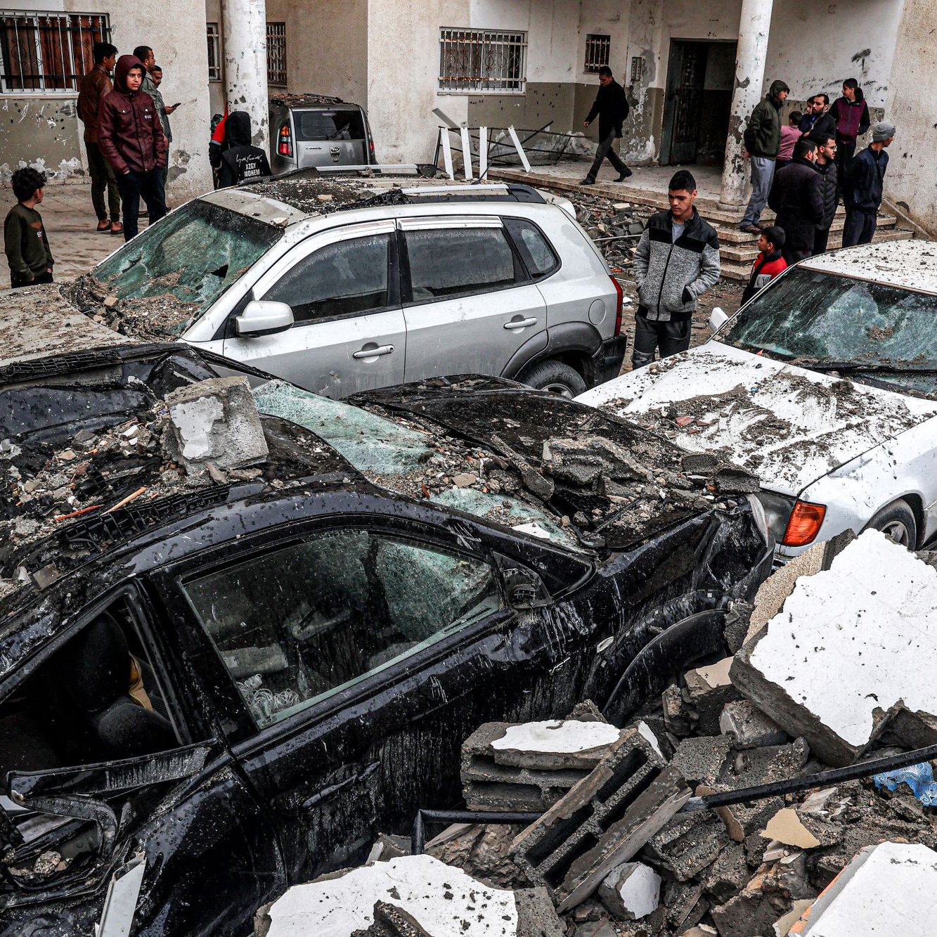 L'immagine mostra una scena di devastazione dopo un evento disastroso, probabilmente un terremoto. Si possono vedere diverse auto danneggiate, alcune sepolte sotto macerie e detriti. Il contesto è urbano, con edifici visibilmente danneggiati in fondo. Ci sono anche persone nelle vicinanze, che osservano la situazione, alcune sembrano preoccupate o increduli di fronte ai danni. La scena trasmette un forte senso di shock e di difficoltà.