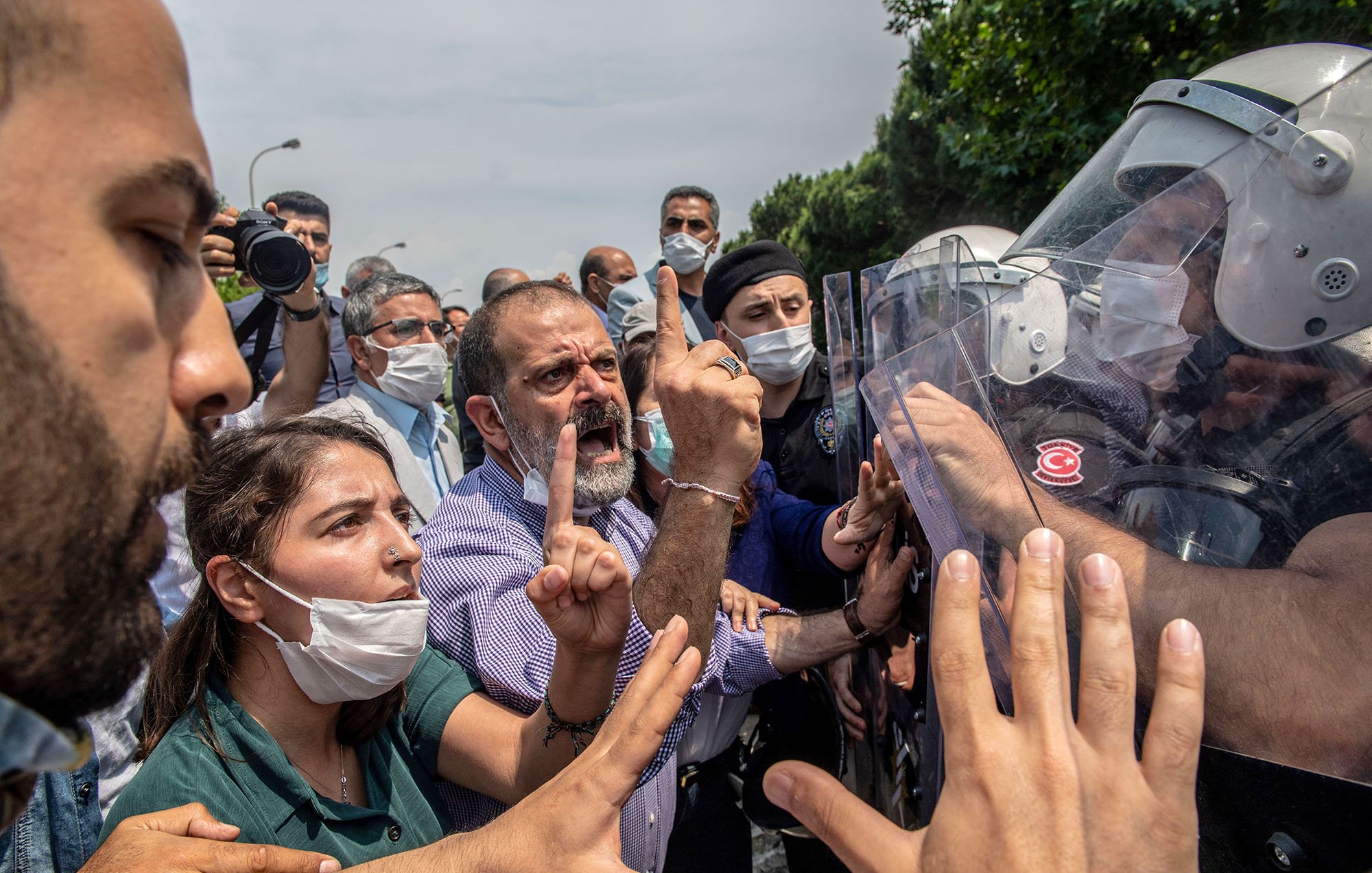 تظهر الصورة محتجين يتجمعون في مكان عام، حيث يبدو أن هناك توترًا في الأجواء. بعض الأشخاص يحملون لافتات، بينما يتواجد عدد من رجال الشرطة الذين يرتدون خوذات ودروع لمواجهة الحشد. يبدو أن هناك تفاعلًا حادًا بين المتظاهرين ورجال الأمن، مع تعبيرات وجهية تشير إلى الغضب والاحتجاج. بعض الأشخاص يرتدون أقنعة للوجه، مما يدل على احتمال وجود تدابير صحية. الأشجار والخضرة، تشير إلى أن الموقع في منطقة حضرية.