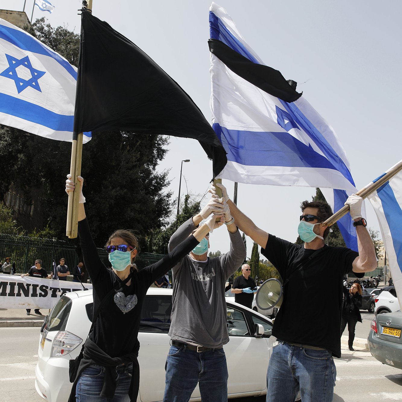 L'image montre un groupe de personnes en train de manifester. Elles tiennent des drapeaux, y compris le drapeau israélien et un autre drapeau aux couleurs noire et blanche. Tous les participants portent des masques sur le visage, ce qui suggère qu'ils prennent des précautions sanitaires. En arrière-plan, on peut voir une banderole qui semble contenir un message critique envers le gouvernement. L'atmosphère semble être celle d'une protestation pacifique.