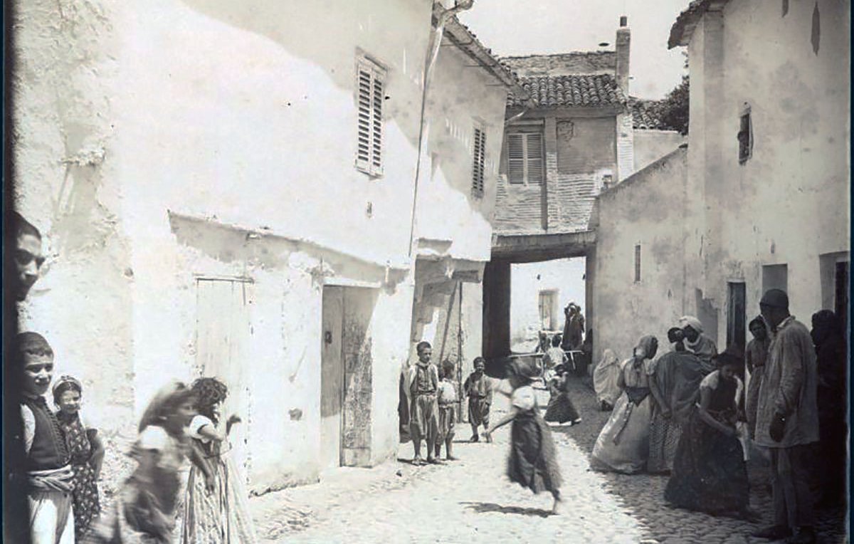 L'image représente une scène de rue dans un quartier de Constantine, en Algérie. On y voit des maisons blanches avec des volets, et une ruelle pavée qui semble étroite. Plusieurs personnes, y compris des enfants, sont visibles, certaines marchant et d'autres se tenant debout. Les vêtements des habitants semblent traditionnels, reflet d'une époque passée. L'atmosphère générale évoque une vie quotidienne animée dans un contexte historique.