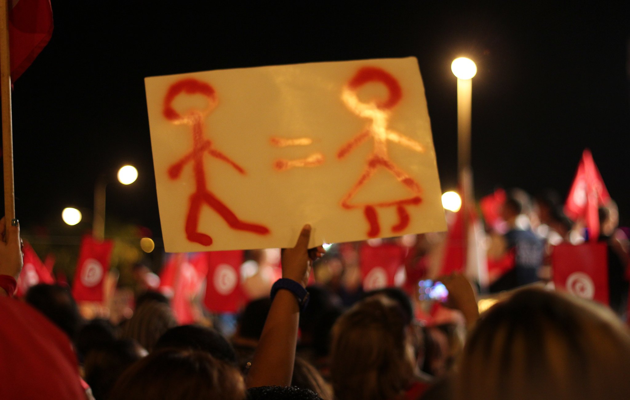 The image shows a night scene of a demonstration or rally, where a person is holding up a sign. The sign features a simplistic representation of a male figure and a female figure, with an equals sign between them, suggesting a message of gender equality. In the background, a crowd can be seen, along with red flags that likely represent a political or social movement. The atmosphere appears to be vibrant and engaged, reflecting a collective message or cause.