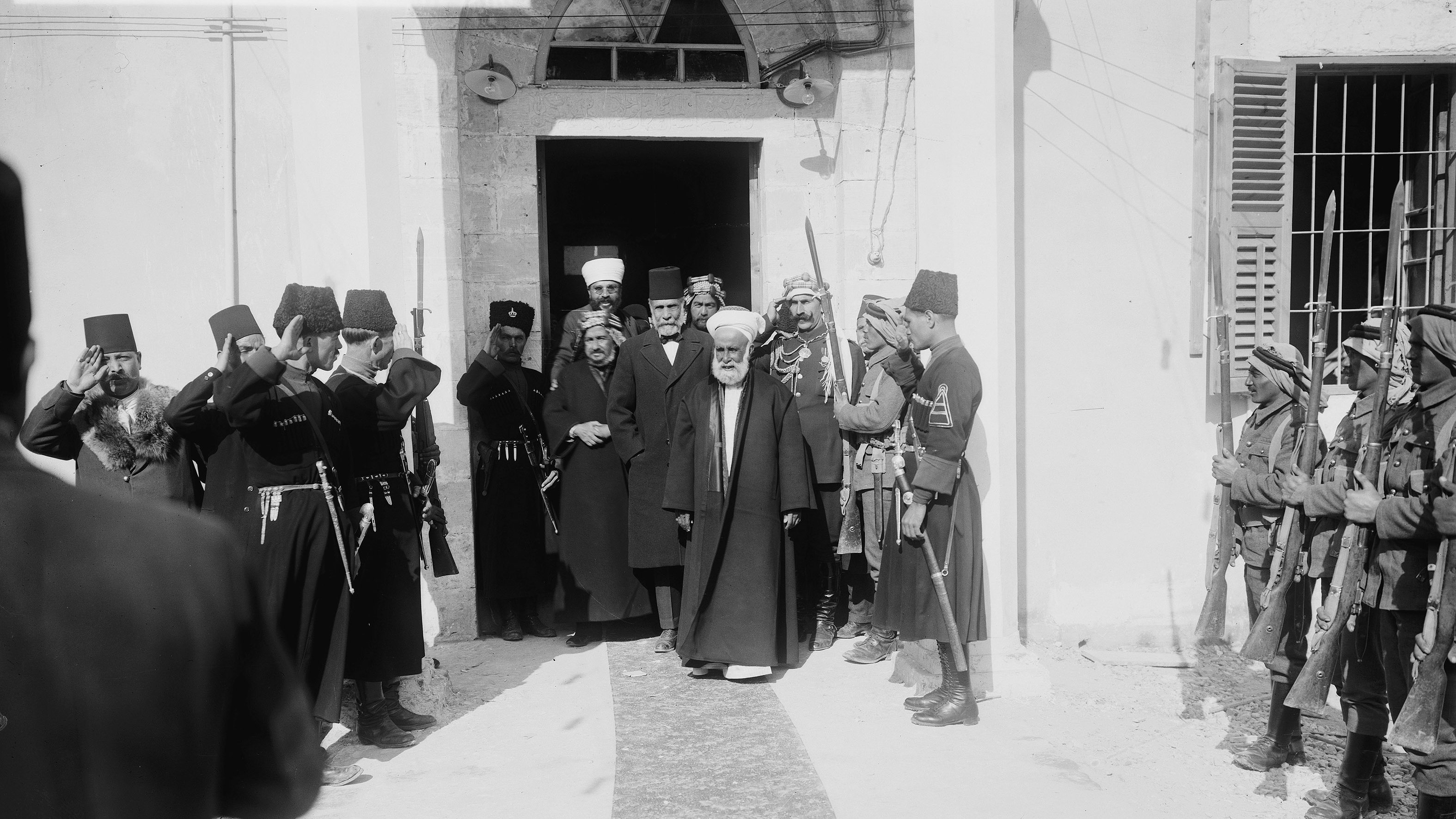در تصویر، گروهی از مردان در یک مکان رسمی ایستادهاند. چند نفر از آنها در لباسهای نظامی و دیگران در لباسهای سنتی دیده میشوند. مردی با چهرهای جدی و کت و شلوار تیره در حال قدم گذاشتن به سمت دوربین است، در حالی که دیگران به او احترام میگذارند و سلام نظامی میدهند. محیط تصویر به نظر میرسد که یک ساختمان تاریخی یا دولتی باشد. فضای کلی نشاندهندهی یک واقعه مهم و رسمی است.