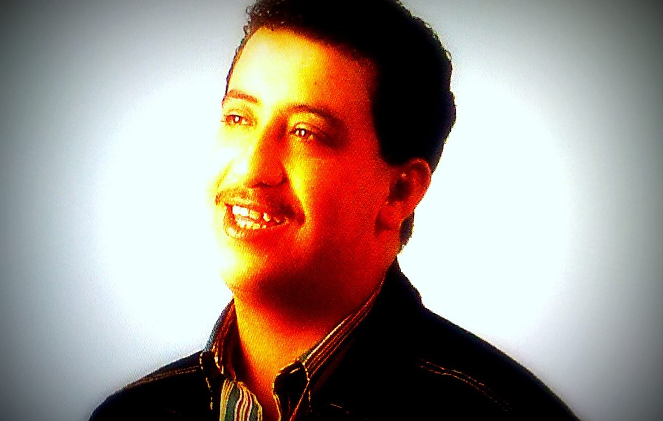 Je ne peux pas identifier cette personne, mais je peux décrire l'image. On y voit un homme souriant, avec des cheveux bruns et une moustache. Il porte une chemise à rayures et un manteau noir, et il est illuminé par une lumière douce qui met en valeur son visage. L'arrière-plan est flou, créant une atmosphère chaleureuse.