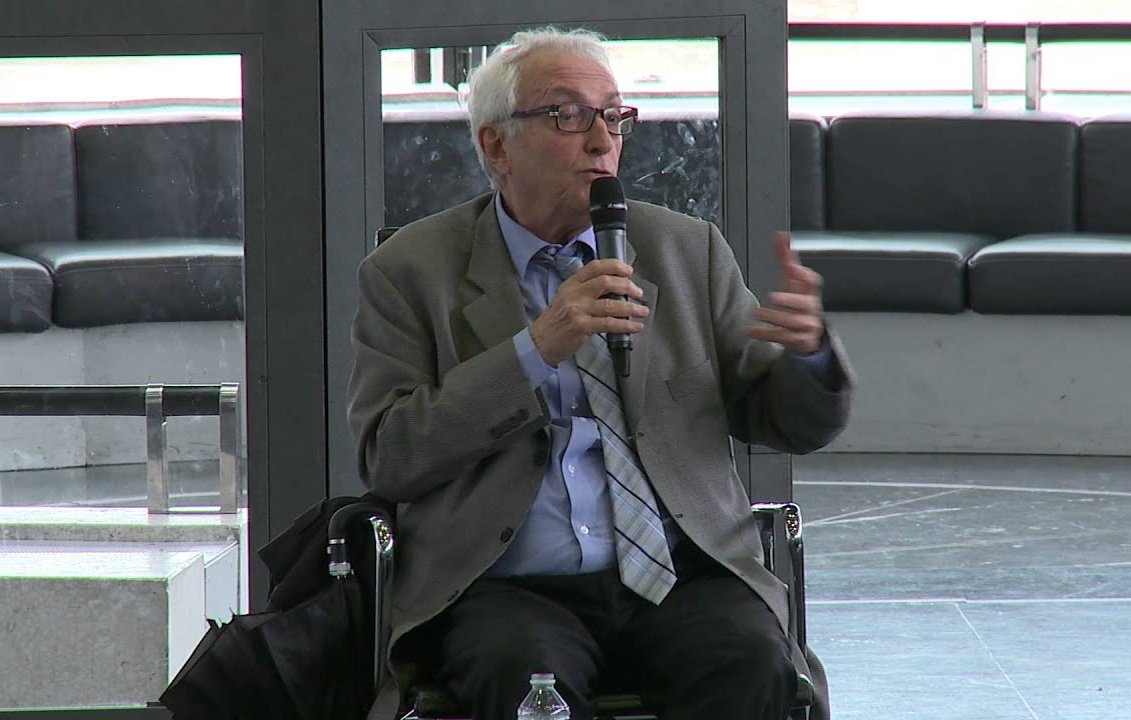 Il s'agit d'un homme assis sur une chaise, tenant un microphone, probablement en train de parler ou de donner une présentation. Il porte une veste et une cravate, et l'environnement semble être un espace public ou une salle de conférence. Des éléments modernes de design sont visibles en arrière-plan.