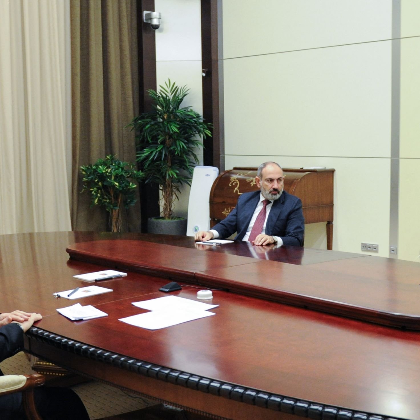 L'image montre une réunion formelle dans une salle ornée. Trois hommes sont assis autour d'une grande table ovale en bois. Au centre, un homme en costume parlant, probablement un chef d'État ou un dirigeant. À sa gauche, un homme au visage sérieux, également en costume. À droite, un autre homme, avec une expression attentive. La pièce est décorée de rideaux et d'une plante d'intérieur, ce qui lui donne un aspect sobre et professionnel.