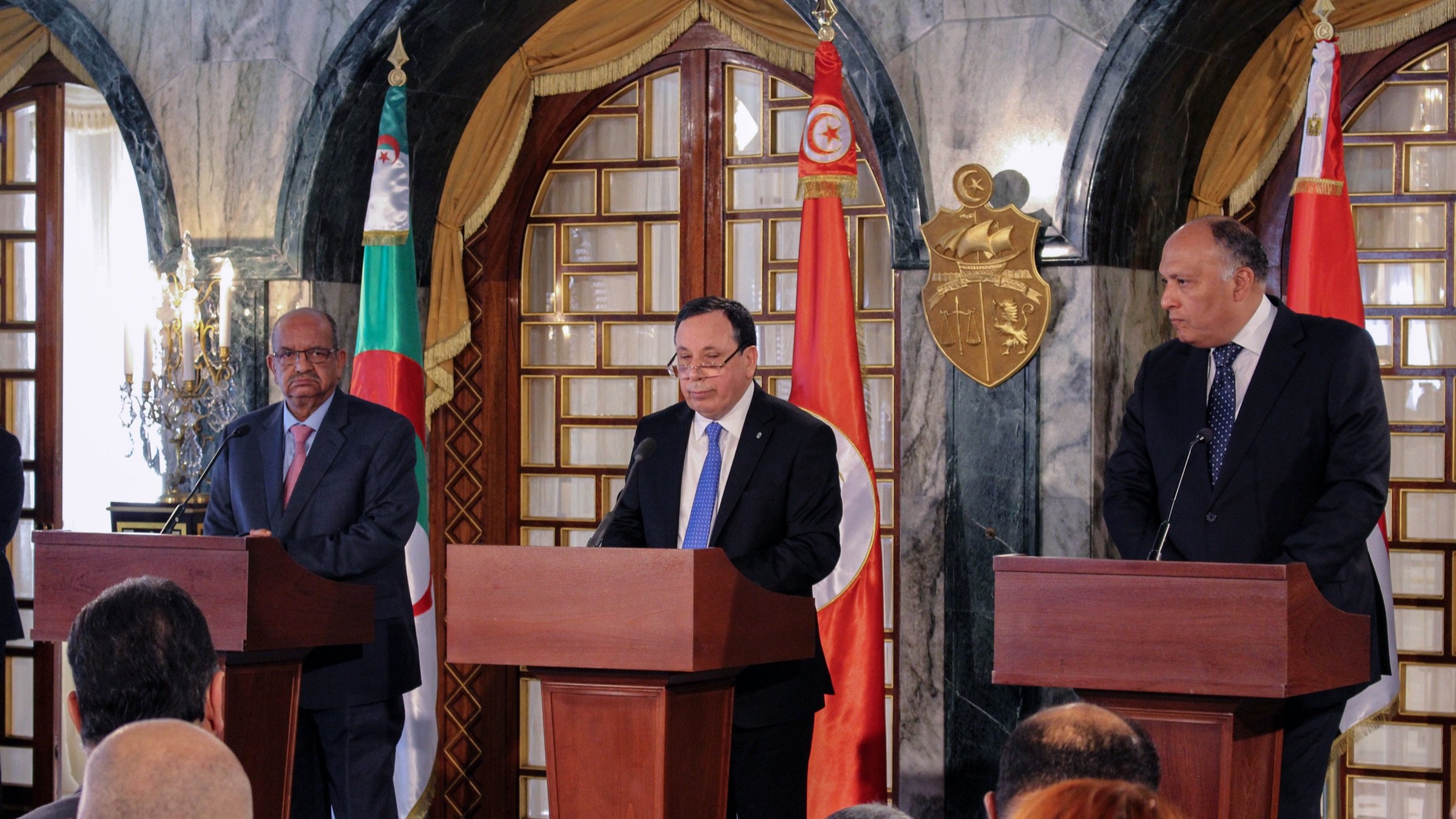 The image depicts a press conference or official meeting involving three individuals standing behind wooden podiums. The man in the center appears to be speaking, while the two flanking him are listening. The backdrop features flags of Algeria and Tunisia, along with a decorative emblem. The setting has an elegant, formal ambiance, likely within a government building. There are several journalists or attendees seated in front, capturing the event.