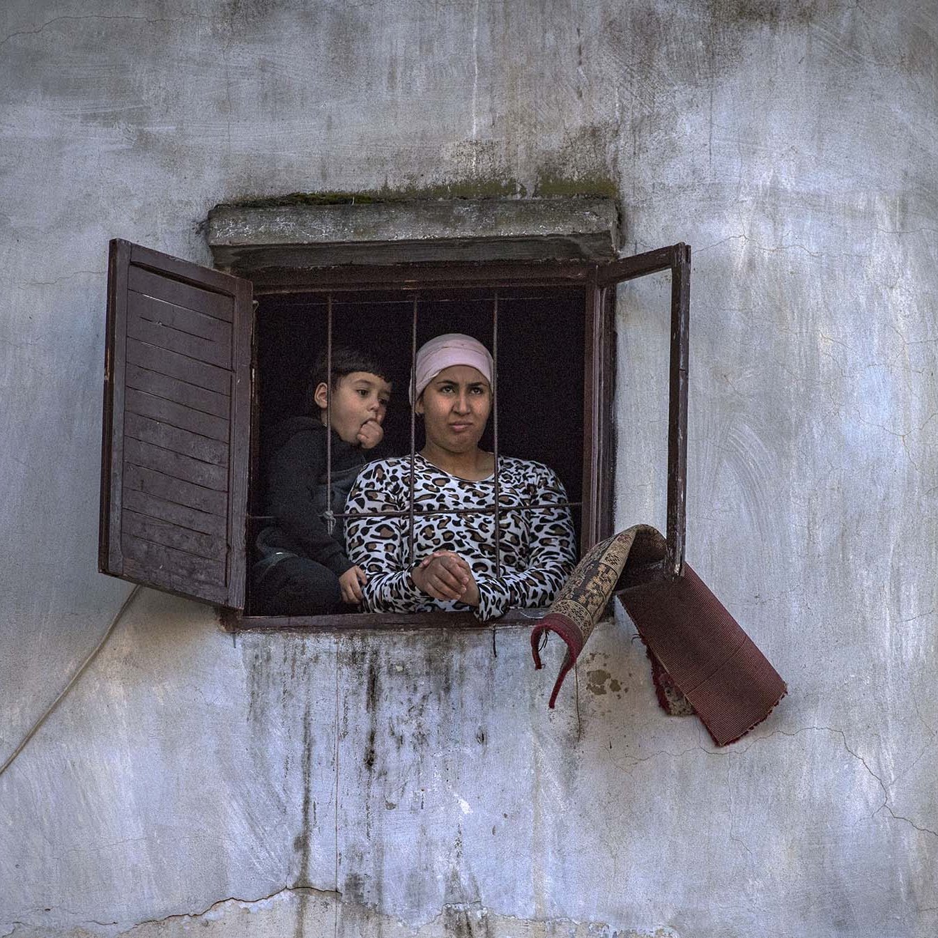 L'image montre deux personnes regardant à travers une fenêtre. Il y a une femme, vêtue d'un haut à motifs, qui semble attentive. À côté d'elle, un enfant, visiblement curieux, se penche légèrement. L'environnement est simple, avec des murs en crépi et une fenêtre aux volets ouverts. L'atmosphère semble réfléchie et calme.