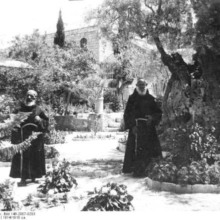L'image montre deux hommes en habits religieux, probablement des moines, se tenant debout dans un jardin. Ils se tiennent près d'un grand arbre, qui semble être un olivier très ancien. L'environnement est rempli de verdure, avec des plantes et des buissons autour. En arrière-plan, on peut apercevoir des murs en pierre, suggérant un cadre historique ou religieux. L'atmosphère semble paisible et sereine.