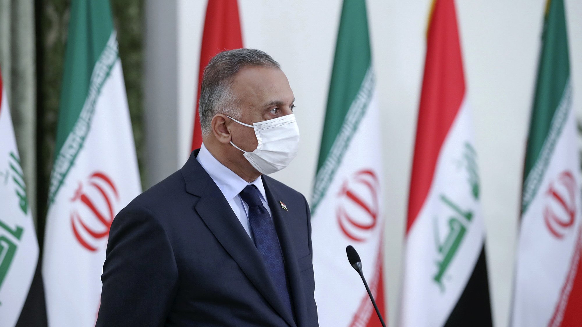 L'image montre un homme en costume, portant un masque facial. Il se tient devant plusieurs drapeaux, dont ceux de l'Iran et de l'Irak. Son expression semble sérieuse, et il semble être en train de participer à une conférence ou une réunion. Le décor est formel, suggérant un cadre politique ou diplomatique.