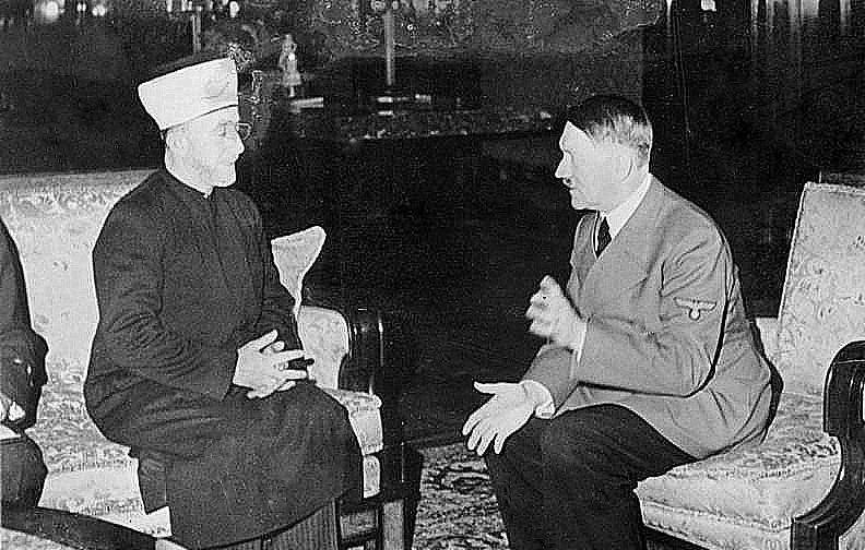 Cette image montre deux hommes assis dans une pièce. L'un d'eux porte un habit traditionnel avec un couvre-chef, tandis que l'autre est vêtu d'un uniforme militaire. Ils semblent être en conversation, gesticulant avec leurs mains. L'environnement est richement décoré, avec des meubles élégants et des éléments de décoration en arrière-plan. L'image évoque un moment de rencontre entre ces deux figures historiques.