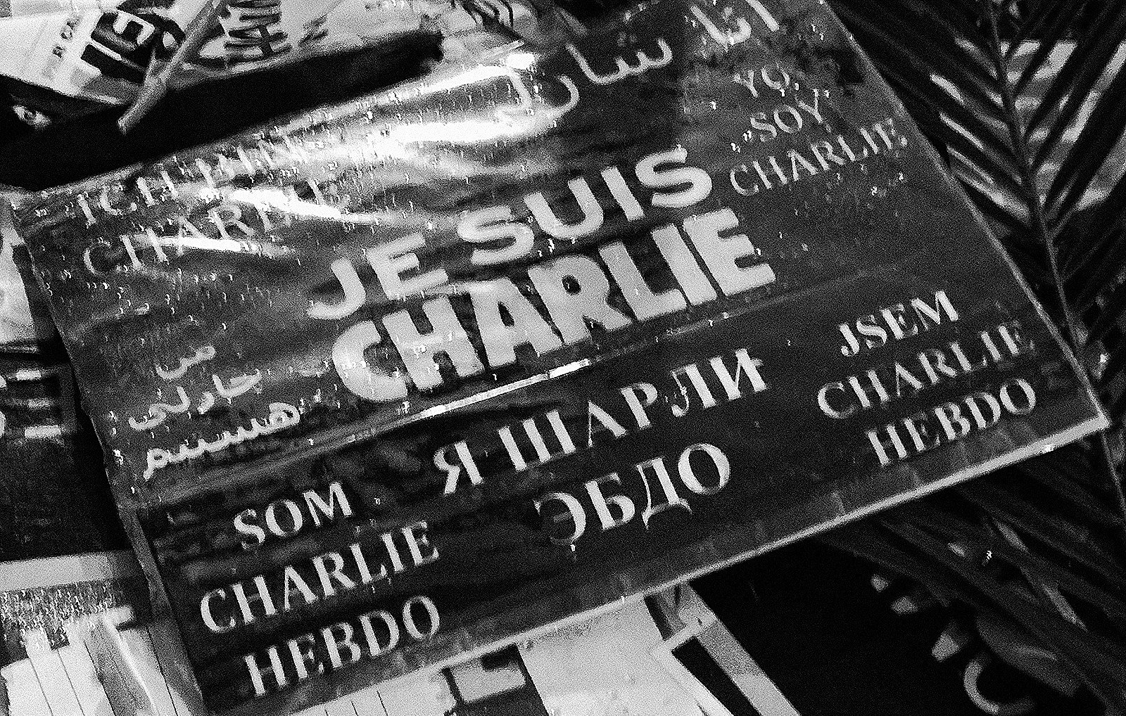 L'image montre une affiche ou un panneau avec l'inscription "JE SUIS CHARLIE" en plusieurs langues. Les mots sont écrits en noir sur fond blanc, et le tout a un aspect qui évoque le soutien et la solidarité, probablement en rapport avec les événements tragiques auxquels cette phrase fait allusion. On peut également voir des éléments environnants qui semblent être des morceaux de papier ou des affiches, ce qui donne un aspect de rassemblement ou de manifestation.