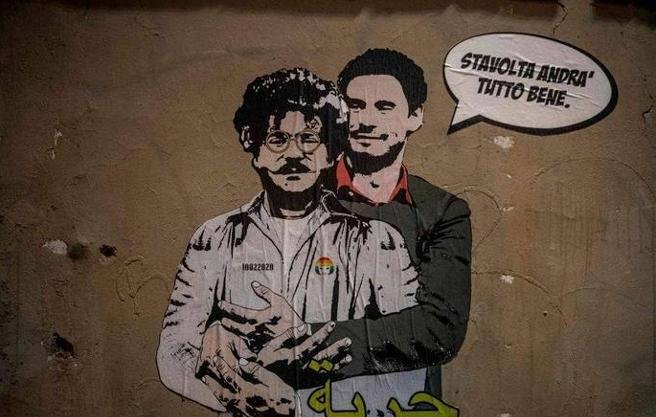 L'immagine mostra un murale che ritrae due figure. Una di esse ha un aspetto distintivo, con occhiali e baffi, mentre l'altra indossa un completo elegante. Sullo sfondo, si può notare un muro di colore neutro, e sopra le figure è presente un testo in italiano che dice "Stai tranquillo, andrà tutto bene". Inoltre, nella parte inferiore della figura è scritta la parola "حرية" (che significa "libertà" in arabo). L'opera sembra trasmettere un messaggio di speranza e solidarietà.