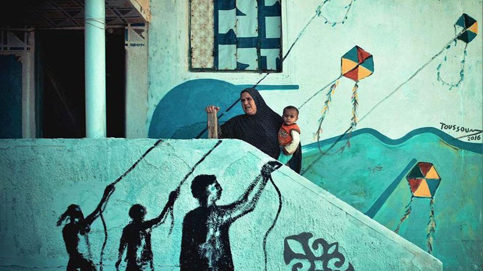 L'image montre une femme, vêtue d'un hijab, tenant un enfant dans ses bras, debout sur une marche d'escalier. En arrière-plan, on peut voir un mur peint avec des motifs colorés et des cerfs-volants. Sur la marche, il y a une fresque représentant des silhouettes de personnes tenant des cerfs-volants, créant un contraste entre l'art mural et la scène réelle. L'ensemble de la composition évoque un sentiment de nostalgie et de connexion entre le passé et le présent.