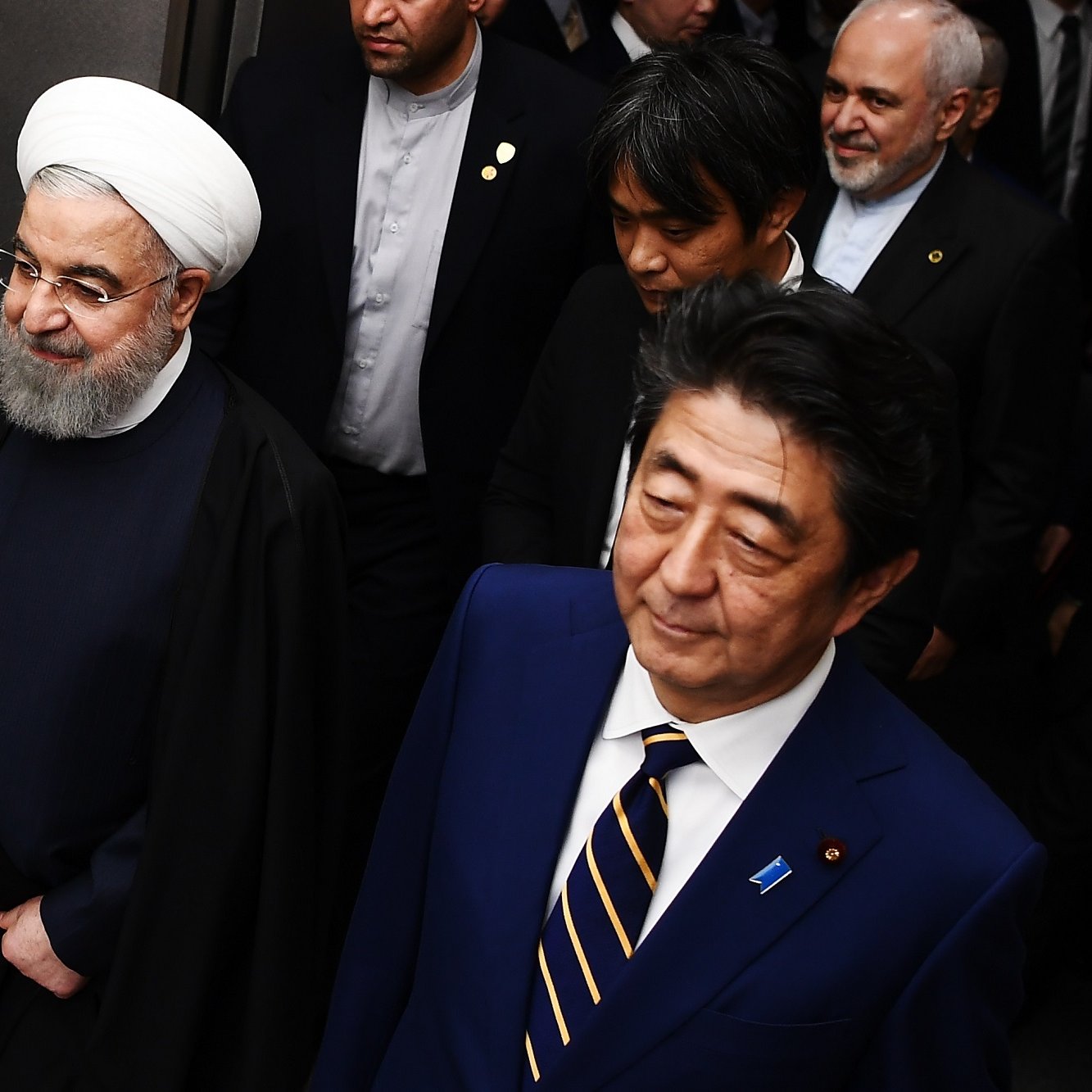 در تصویر، دو مرد در حال پیاده‌روی هستند. یکی از آنها دارای پوشش سنتی ایرانی با عمامه سفید و جبه است، و به نظر می‌رسد که او مقام رسمی و بلندپایه‌ای باشد. فرد دیگر در کنار او، با کت و شلوار رسمی و کراوات، لبخند به لب دارد. در پس‌زمینه، چند نفر دیگر نیز دیده می‌شوند که به نظر می‌رسد همراهان آنها هستند. محیط تصویر ممکن است یک کنفرانس یا جلسه رسمی را نشان دهد.