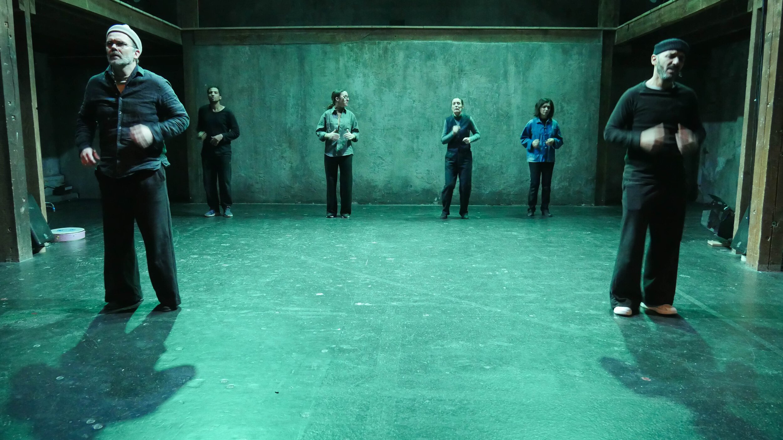 L'image montre un groupe de six personnes sur scène, dans un espace artistique éclairé d'une lumière verte. Elles semblent engagées dans une performance ou une répétition. Les membres du groupe portent des vêtements de différents styles, et se déplacent à divers endroits de la scène. L'arrière-plan est dépouillé, avec des murs en béton, ce qui crée une ambiance minimaliste et contemporaine.