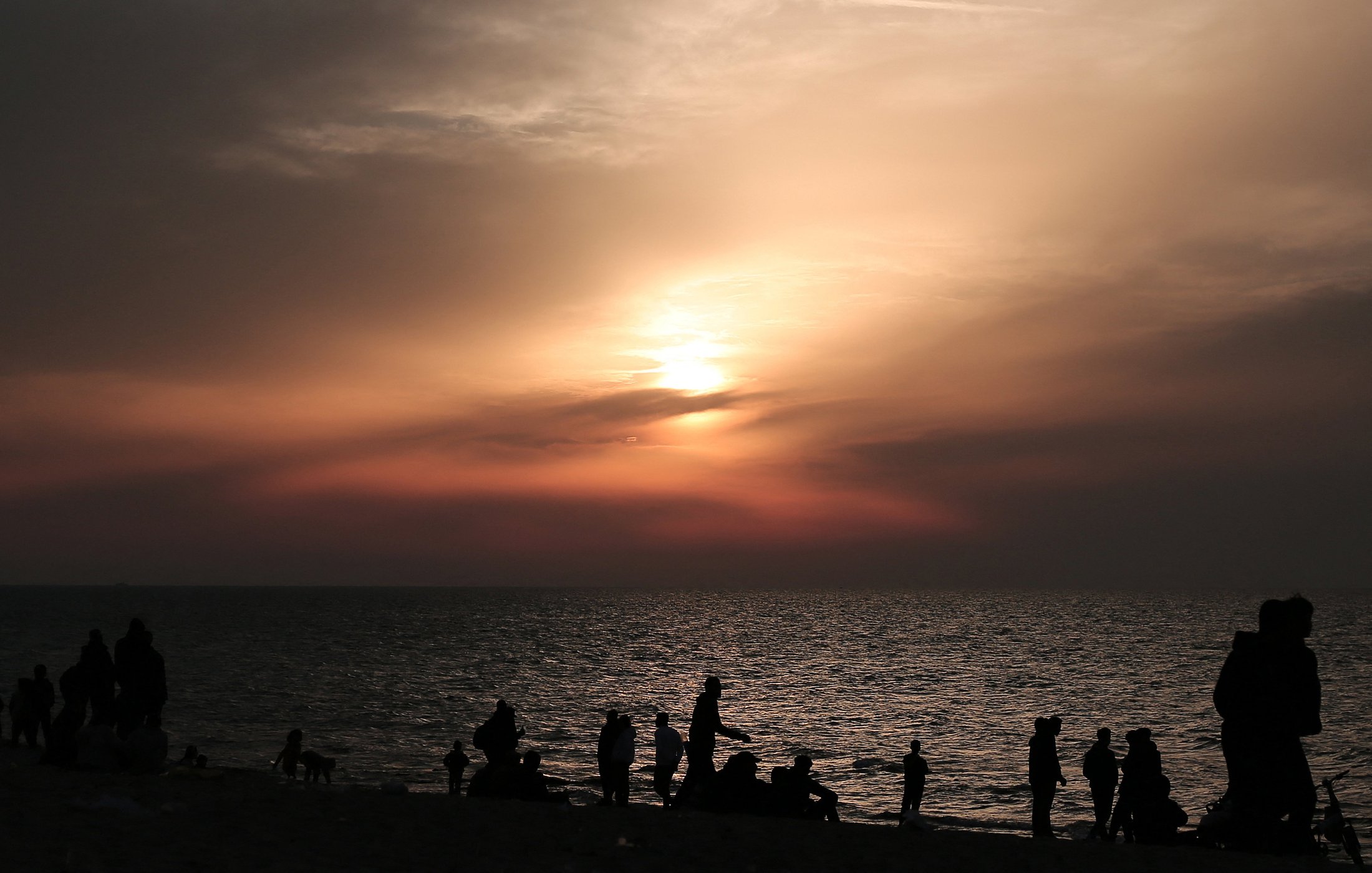The image depicts a beach scene during sunset. In the foreground, there are silhouettes of people walking along the shore, some sitting or standing by the water. The sky is filled with soft hues of orange and pink, blending into darker clouds as the sun sets on the horizon. The water reflects the colors of the sky, creating a tranquil and serene atmosphere. Overall, the scene conveys a sense of calm and natural beauty as day transitions into night.