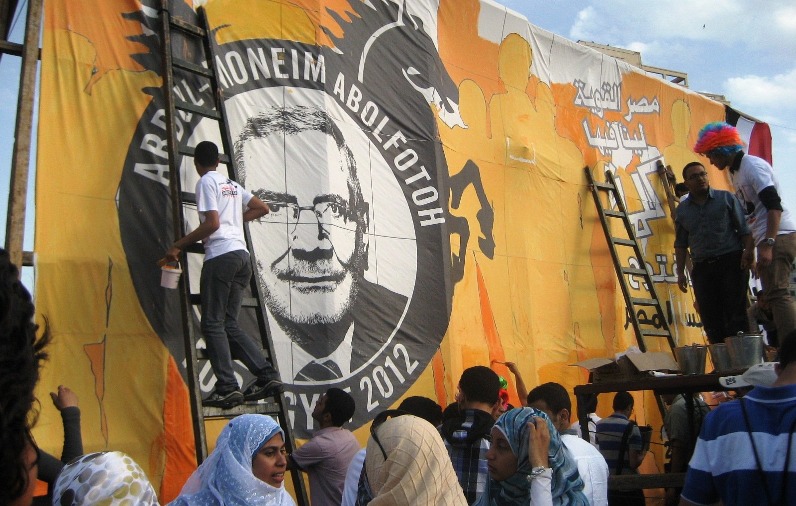 تظهر الصورة مجموعة من الأشخاص يعملون على رسم وإعداد لوحة ضخمة تحمل صورة وشعار أحد المرشحين الانتخابيين، حيث يقوم بعضهم بتلوين اللوحة باستخدام سلالم. الخلفية برتقالية مع تفاصيل أخرى، ويبدو أن الحدث مرتبط بالانتخابات في عام 2012. يظهر أفراد يرتدون ملابس متنوعة، بما في ذلك بعض الأشخاص الذين يرتدون أغطية رأس تحمل ألوان مختلفة. أجواء العمل والاجتهاد تسيطر على المشهد.