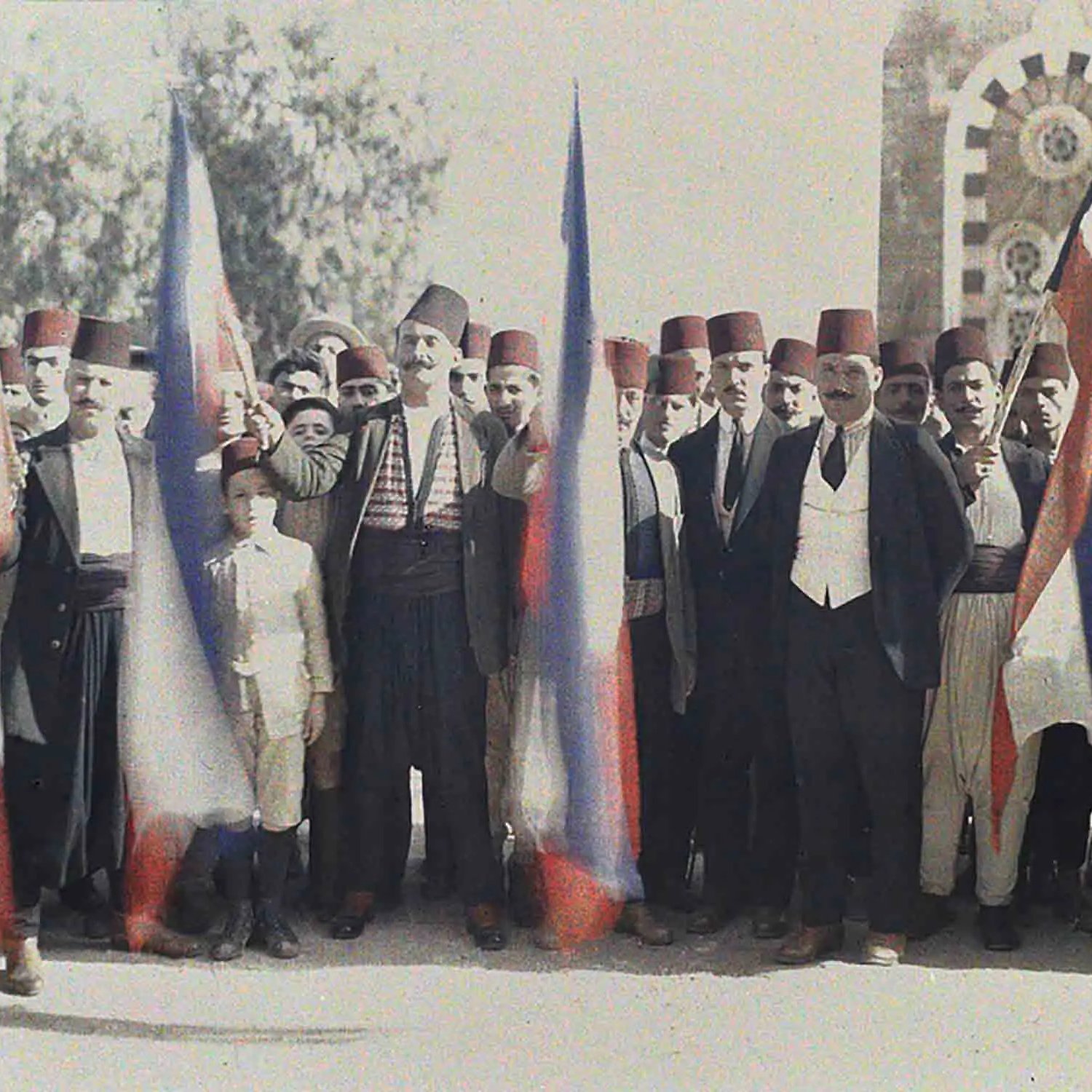 L'image montre un groupe de personnes en tenue traditionnelle, se tenant ensemble avec des drapeaux russes. Les hommes portent des fez et divers vêtements folkloriques, avec un jeune garçon au centre. L'arrière-plan présente une architecture qui semble historique, peut-être un bâtiment symbolique. L'atmosphère suggère un moment de célébration ou de rassemblement associé à une certaine fierté nationale.