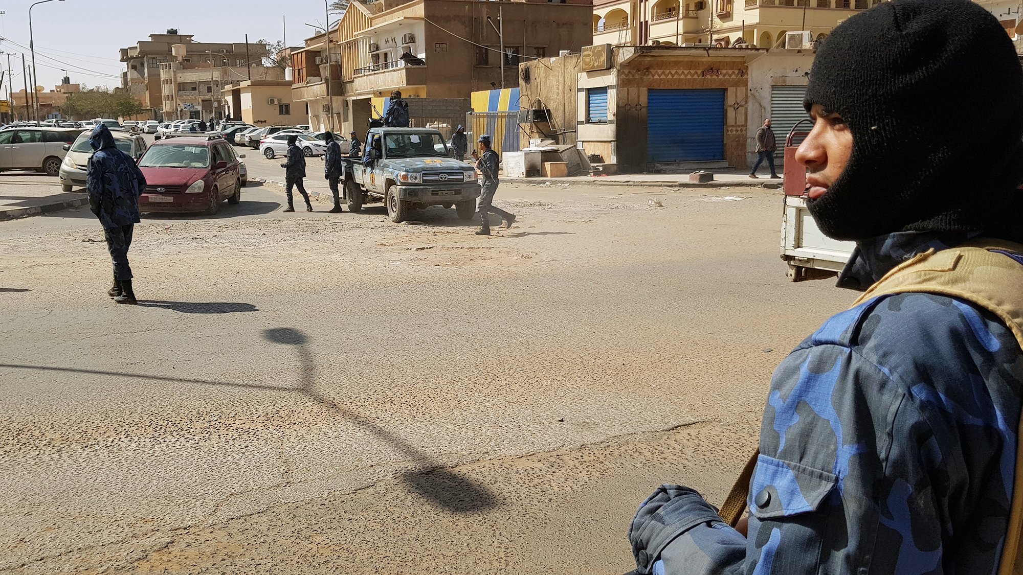L'immagine mostra una scena di una zona urbana, presumibilmente in un contesto di sicurezza elevata. In primo piano, un uomo indossa un berretto e abbigliamento camuffato, mentre tiene un'arma. Sullo sfondo si possono vedere altri uomini in uniformi simili che si muovono in un'area in cui ci sono veicoli parcheggiati. L'architettura circostante appare tipica di una città, con edifici vari e attività commerciali. La luce del giorno illumina la scena, creando ombre ben definite.