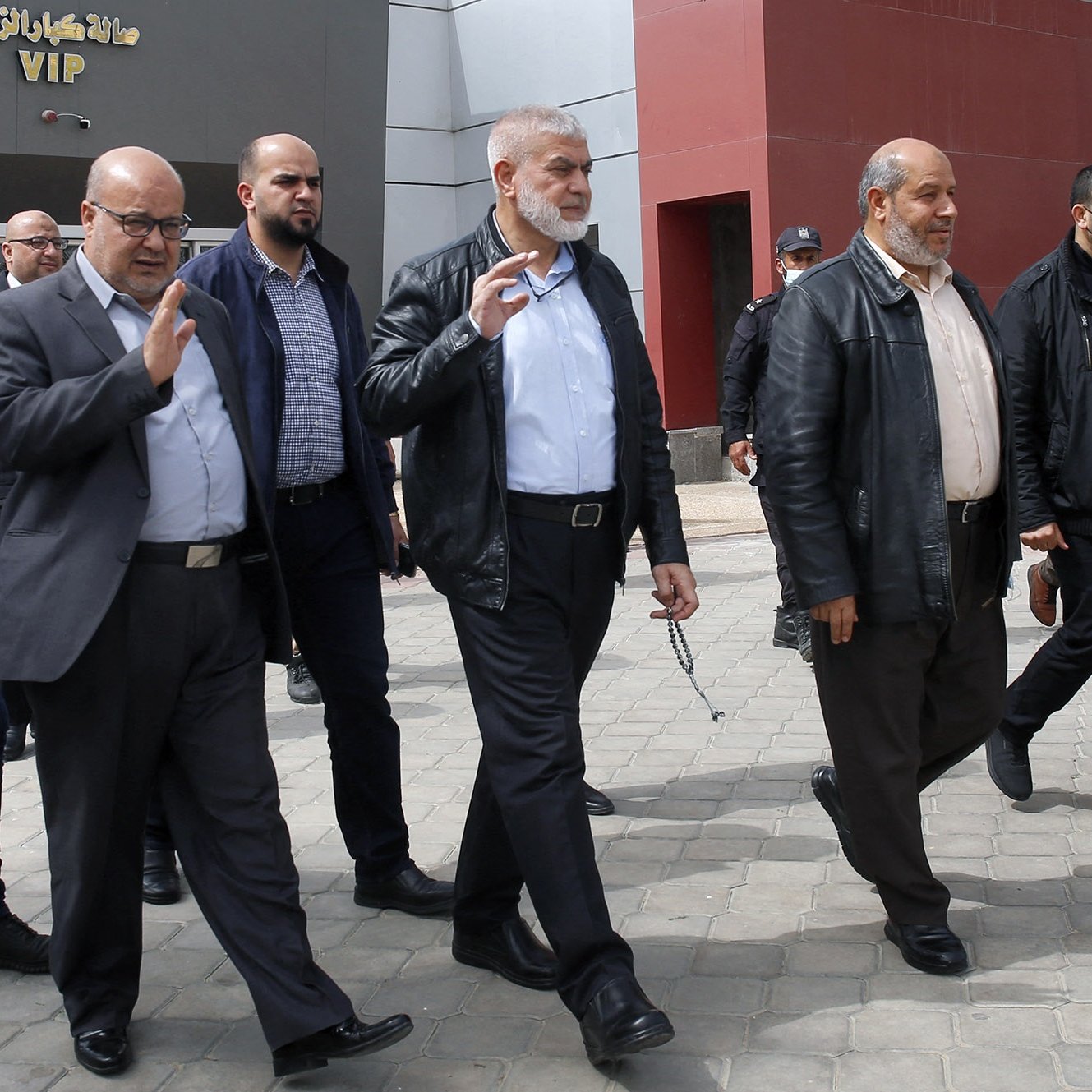 Nell'immagine si vede un gruppo di uomini che camminano insieme all'aperto. Alcuni di loro indossano giacche di pelle e altri abiti formali. Alcuni uomini indossano mascherine, mentre altri no. Sembrano impegnati in una conversazione e uno di loro alza la mano in un gesto di saluto. Sullo sfondo si intravedono strutture architettoniche moderne e una pavimentazione in pietra. L'atmosfera sembra formale e organizzata.