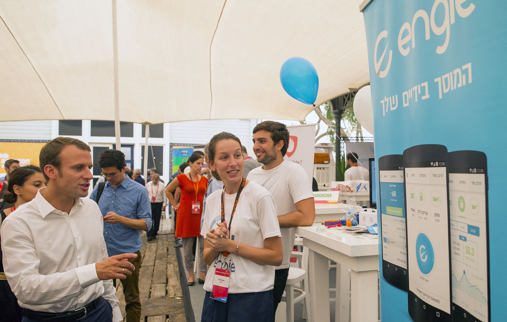 تظهر الصورة مشهداً في حدث أو معرض، حيث يتواجد عدد من الأشخاص تحت خيمة. يتمركز شخص يرتدي قميصاً أبيض، يتحدث مع شخص آخر يبدو أنه زائر. خلفهم، توجد لافتة تحمل شعار "engie" مع صور توضيحية لواجهة تطبيقات على الهواتف. يظهر في الصورة أيضاً عدد من الأشخاص الآخرين في الخلفية، وبعضهم يرتدي ملابس تحمل شارات. الجو يبدو غير رسمي ومليئاً بالنشاط.