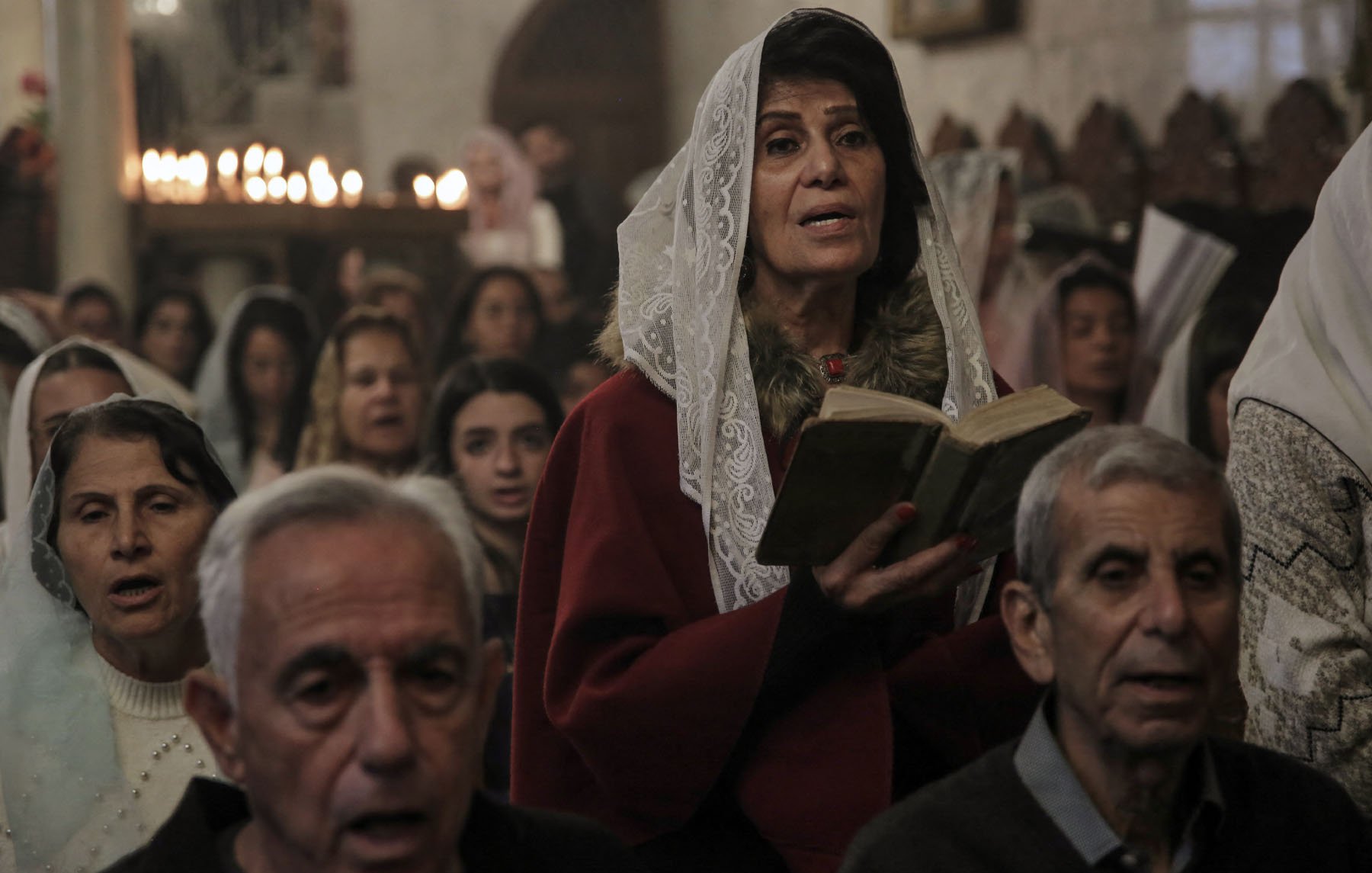 L'immagine mostra un gruppo di persone durante una cerimonia, probabilmente in una chiesa. Una donna al centro, che indossa un velo bianco e un abito rosso, tiene in mano un libro e sembra partecipare attivamente alla celebrazione. Intorno a lei ci sono altre persone, alcune delle quali appaiono in preghiera o cantando. L'atmosfera è solenne, con un gioco di luci morbide e candele accese sullo sfondo che creano un'illuminazione calda e accogliente.