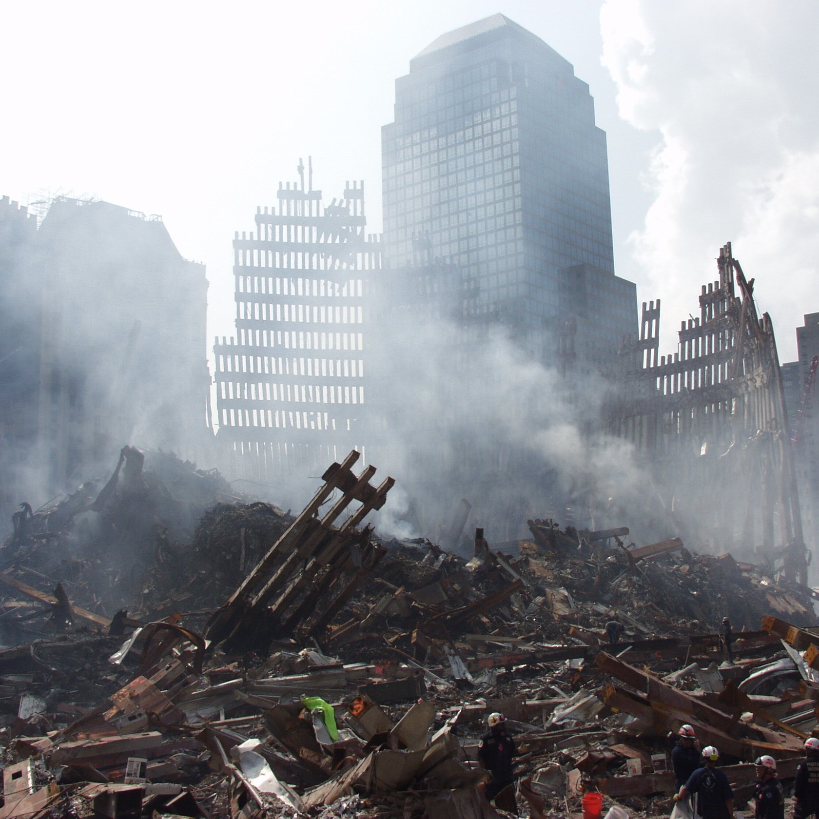 L'image montre un paysage urbain dévasté, avec des débris de bâtiments effondrés et des structures en ruines. De la fumée s'élève dans l'air, créant une atmosphère sombre et chaotique. On peut voir des restes de structures métalliques, avec des morceaux de béton éparpillés sur le sol. L'arrière-plan présente des gratte-ciel partiellement visibles, suggérant une zone qui a subi des dommages significatifs. La scène évoque un sentiment de destruction et de désolation.