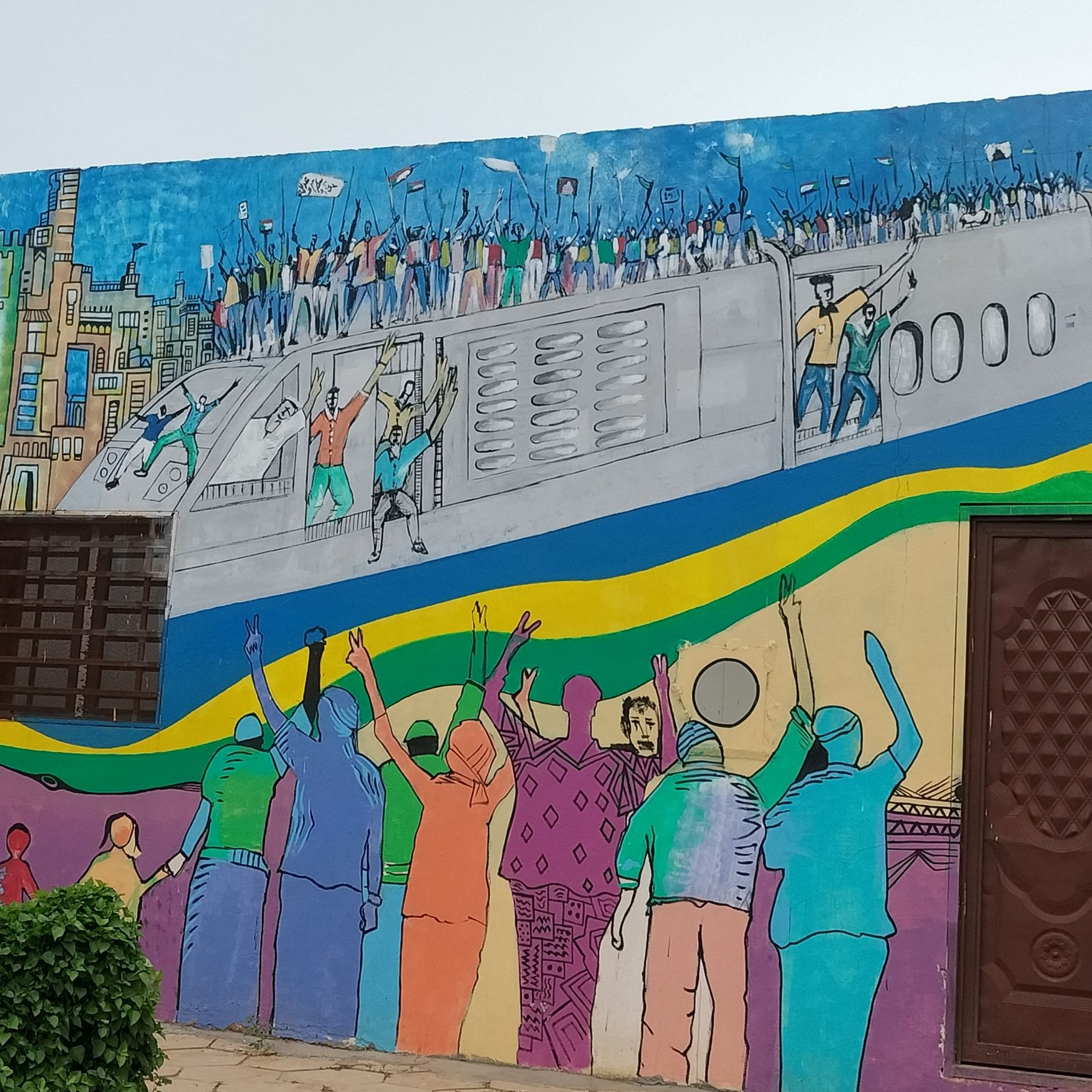 L'image montre une fresque murale colorée qui illustre une scène dynamique. On y voit des personnes célébrant et manifestant, les bras levés, sur un fond représentant un avion et une ville. Les couleurs vives ajoutent une énergie palpable à la scène, reflétant un sentiment de joie ou de solidarité. Des silhouettes de différents personnages sont visibles, et l'ensemble de l'œuvre évoque un message de rassemblement et d'espoir.
