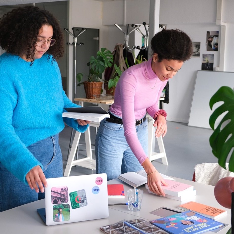 L'image montre deux femmes dans un environnement de travail créatif. L'une d'elles, vêtue d'un pull bleu, semble arranger des choses sur une table. L'autre femme, en pull rose, est en train de manipuler des livres ou des documents. Sur la table, il y a un ordinateur portable, des livres et quelques accessoires de bureau. L'espace est lumineux et décoré de plantes, ce qui contribue à une ambiance agréable et inspirante.