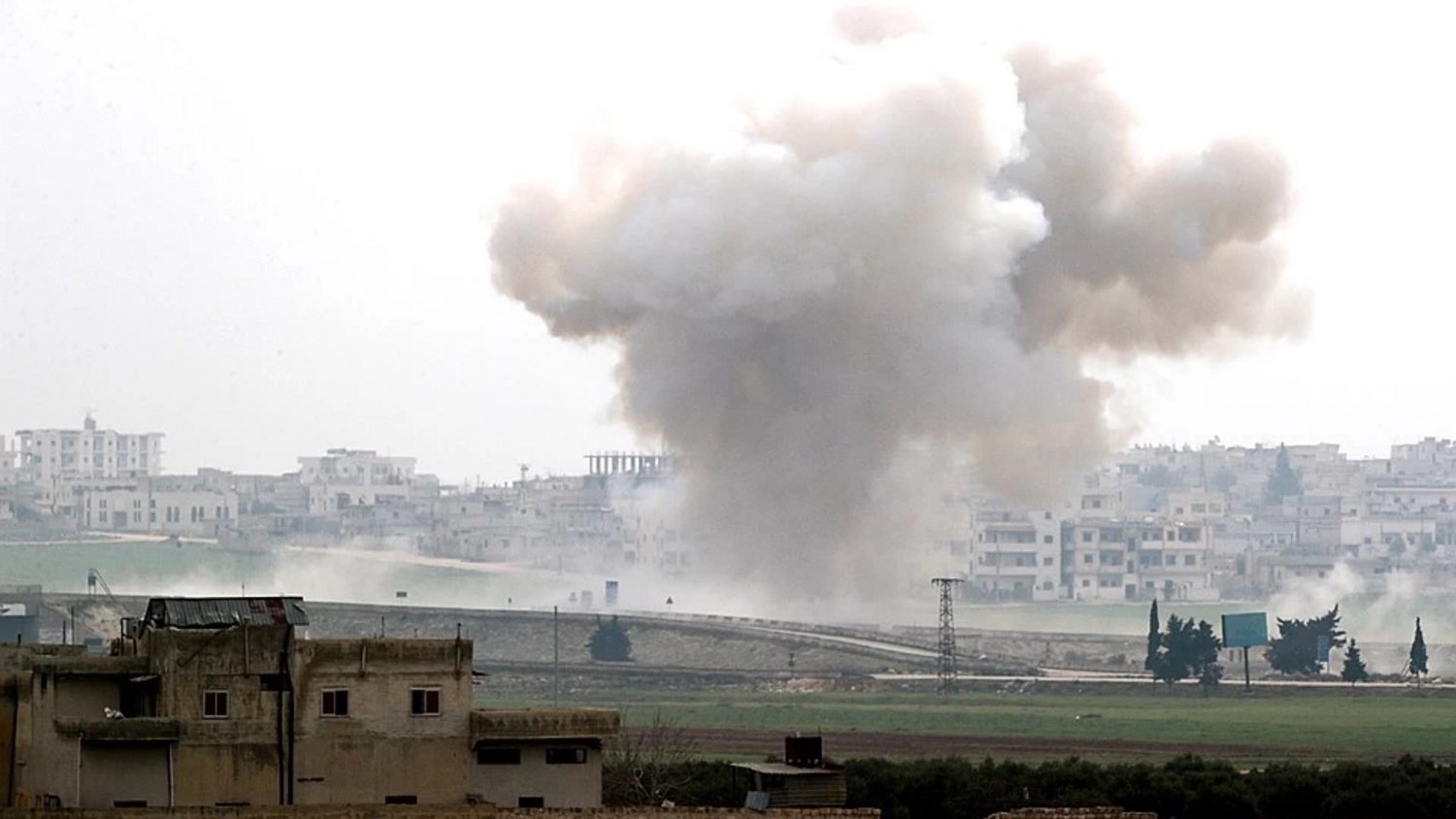L'image montre un paysage de guerre, avec une explosion qui génère un nuage de fumée sombre s'élevant dans le ciel. Au premier plan, on peut apercevoir des bâtiments endommagés, tandis qu'à l'arrière-plan, une ville est visible, probablement touchée par le conflit. L'atmosphère dégage une impression de destruction et de chaos, illustrant les conséquences d'une situation de conflit armé.