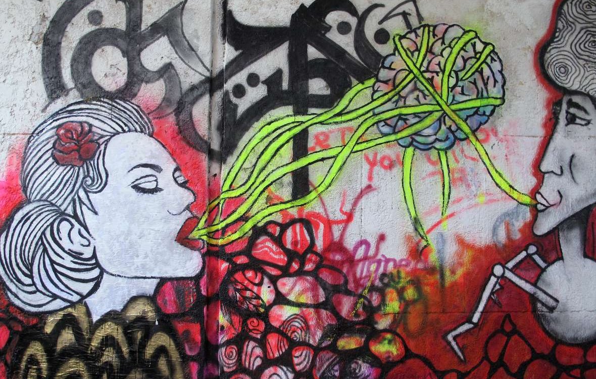 L'image montre une œuvre de street art colorée sur un mur. À gauche, il y a le portrait d'une femme avec un rose dans les cheveux, qui semble "parler" ou "chanter". À droite, un homme est représenté, affichant une expression sérieuse. Entre les deux, des lignes vertes émanent de la tête de la femme, semblant évoquer une connexion ou un échange de pensées, illustrées par un cerveau stylisé en arrière-plan. L'ensemble est dominé par des couleurs vives et des motifs abstraits, créant une atmosphère vibrante et dynamique.