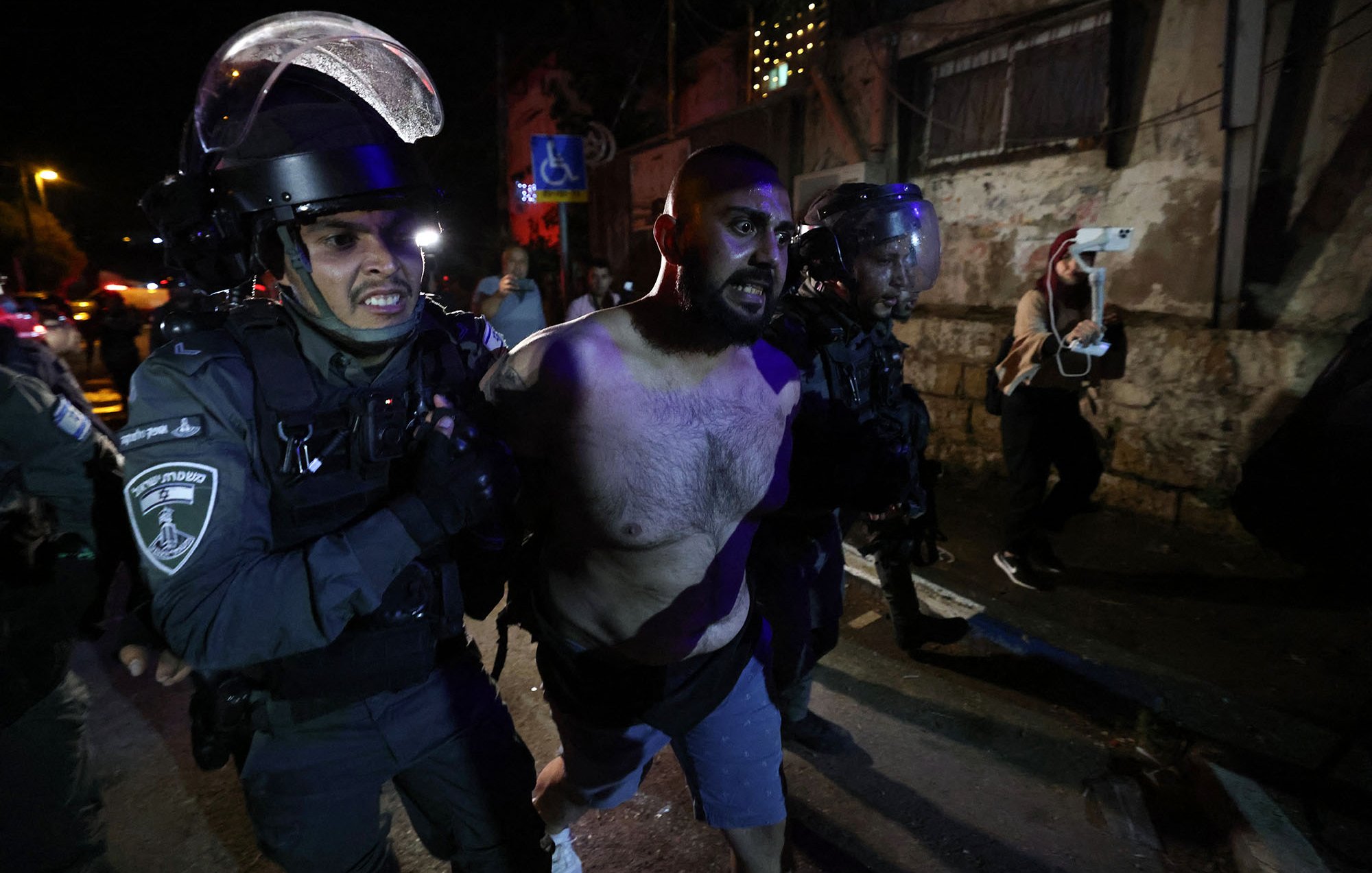 L'image montre des policiers en uniforme, portant des casques et des protections, en train de maîtriser un homme. L'homme est torse nu et semble être en train d'être arrêté. L'environnement nocturne, avec des lumières et des bâtiments en arrière-plan, suggère une situation de tension, potentiellement liée à des manifestations ou des troubles civils. Les expressions et la posture des policiers indiquent une approche ferme envers la situation.
