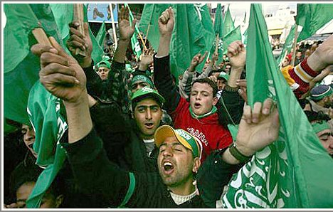 L'image montre une foule de personnes brandissant des drapeaux verts, manifestant de manière énergique. On peut voir des groupes de jeunes hommes exprimer leur enthousiasme, certains chantant ou criant. L'atmosphère semble festive, avec des expressions de joie et de solidarité parmi les participants. Les vêtements et accessoires des manifestants reflètent une allure colorée, souvent associée à des symboles politiques ou culturels.