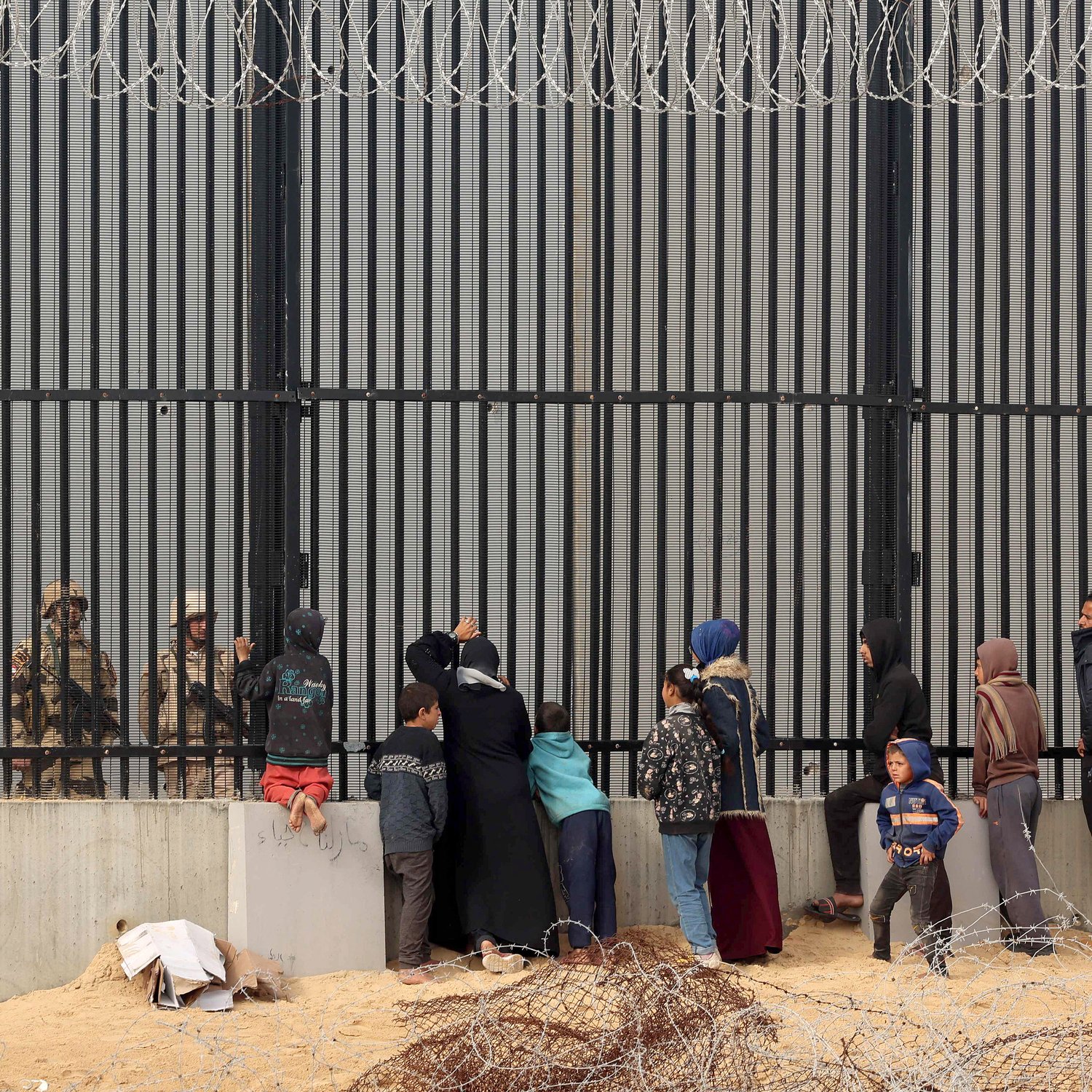L'image montre un groupe de personnes se tenant devant une grande clôture métallique, probablement une barrière de sécurité. De l'autre côté, des soldats en uniforme semblent observer le groupe. Les personnes sont composées de familles et d'enfants, certains d'eux essaient de communiquer ou de se rapprocher de la clôture. Le cadre est marqué par un environnement désertique, avec des barbelés visibles au sol. L'atmosphère semble chargée d'émotion, soulignant une séparation entre les civils et les forces de sécurité.
