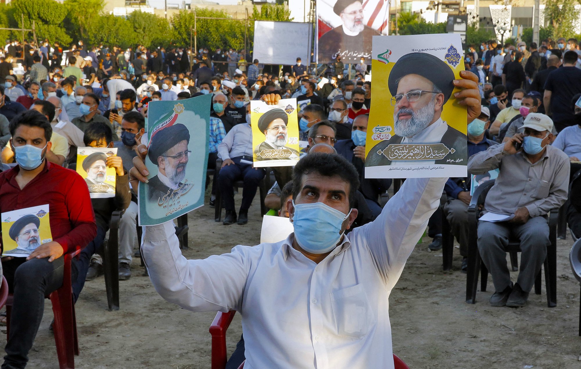 The image shows a large crowd of people seated outdoors, wearing masks. Many attendees are holding posters or pictures of a man, who appears to be a political figure, indicating support for him. The setting looks like a public gathering or rally, with trees in the background and banners visible. The mood appears to be one of engagement and solidarity among the participants.